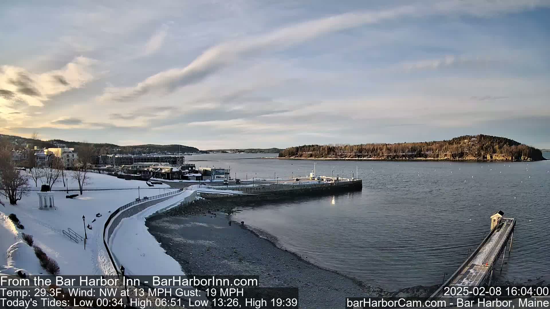 A snow-covered waterfront town with a partially frozen harbor is seen under a partly cloudy sky.