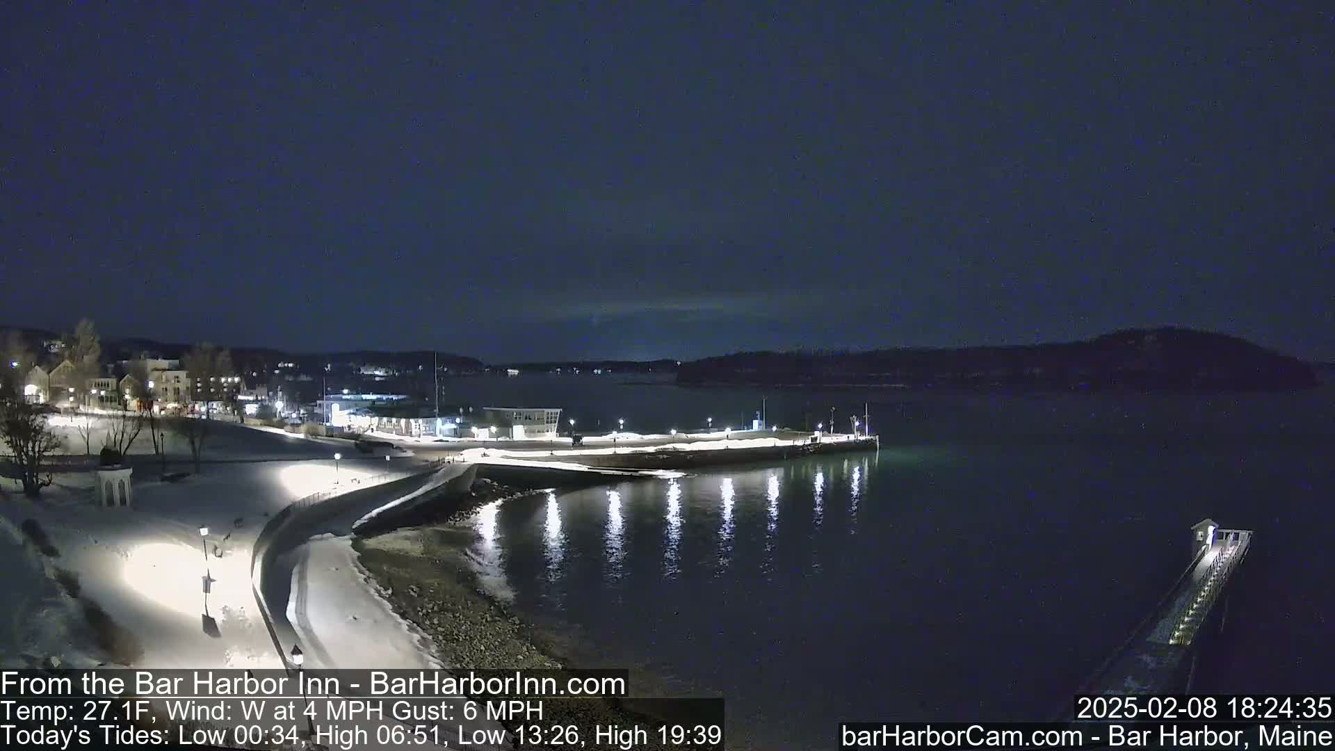 A nighttime view of a snow-covered waterfront town with a calm body of water, illuminated by lights along a shoreline path.