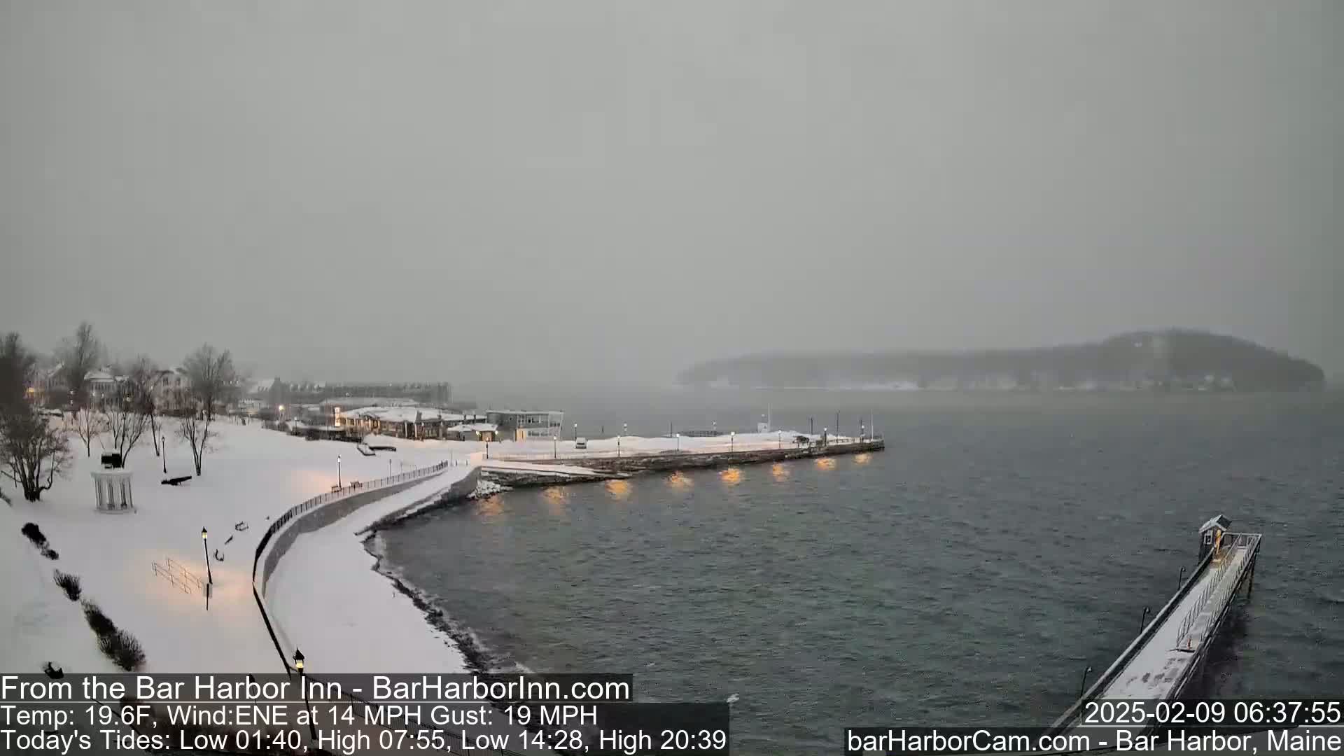 A snow-covered waterfront scene shows a snow-covered walkway and buildings next to a partially frozen body of water, with a pier extending into the water under a gray, overcast sky.
