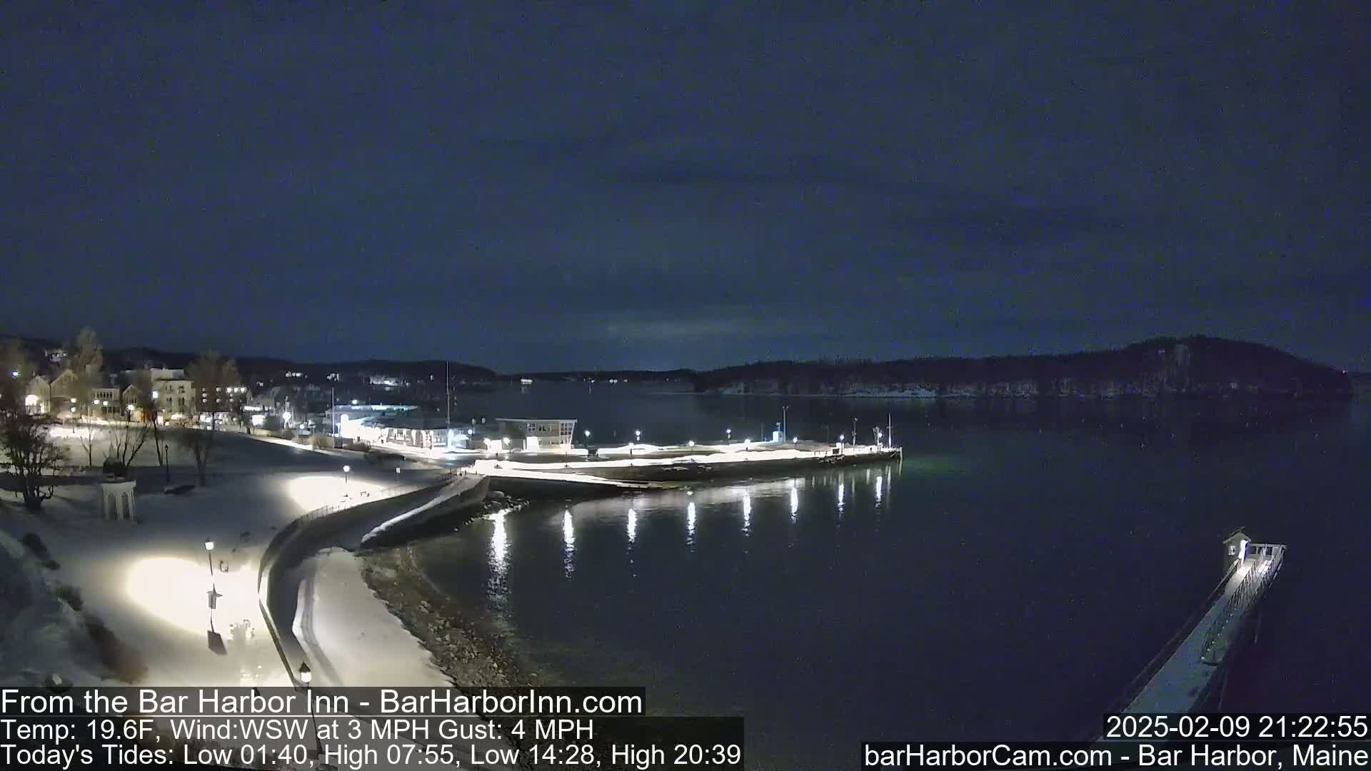 A nighttime view of a snow-covered waterfront town, with calm water reflecting lights from buildings and a pier extending into the bay under a dark, clear sky.