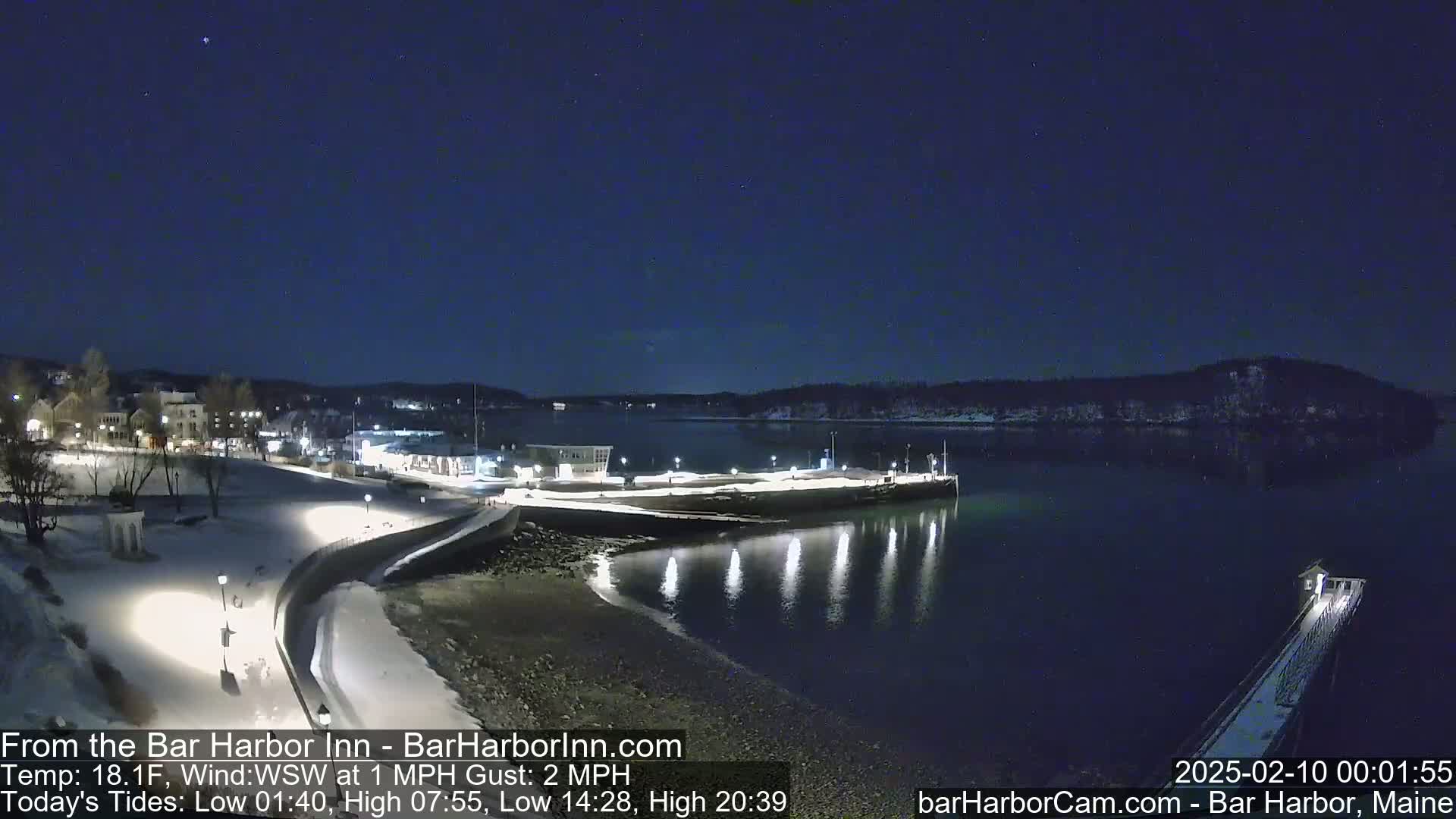 A nighttime view of a snow-covered harbor town with calm water reflecting lights, under a clear, dark sky.