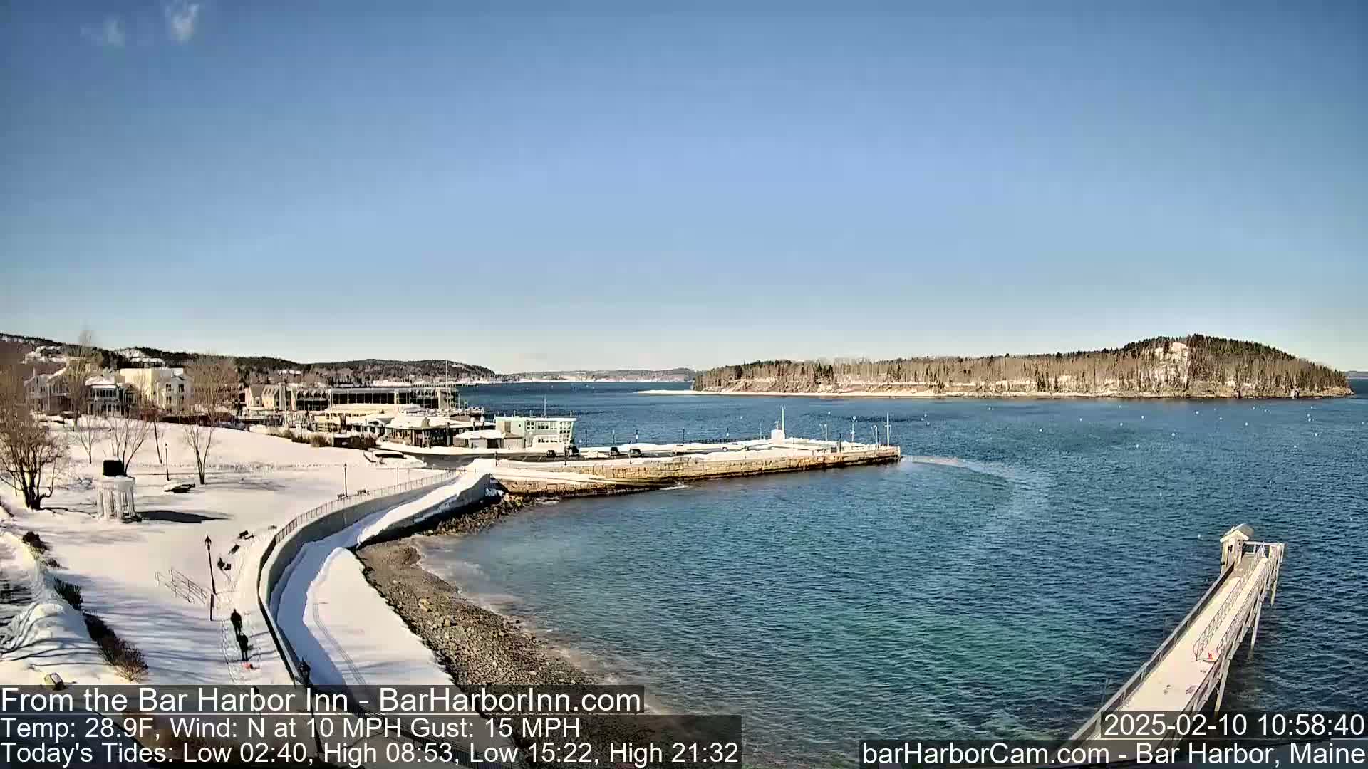 A snow-covered waterfront scene with buildings, a partially frozen harbor, and a long pier extends into the calm, blue water under a clear, sunny sky.