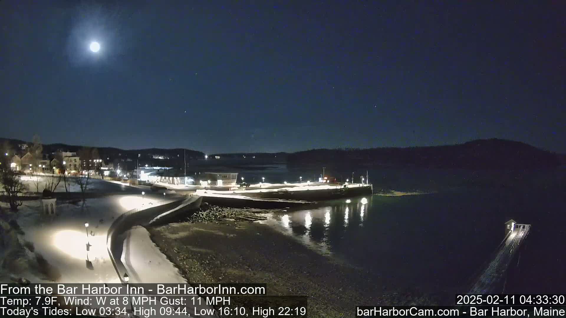 A brightly lit waterfront town is seen under a nighttime sky with a full moon, and a light dusting of snow is present on the ground.