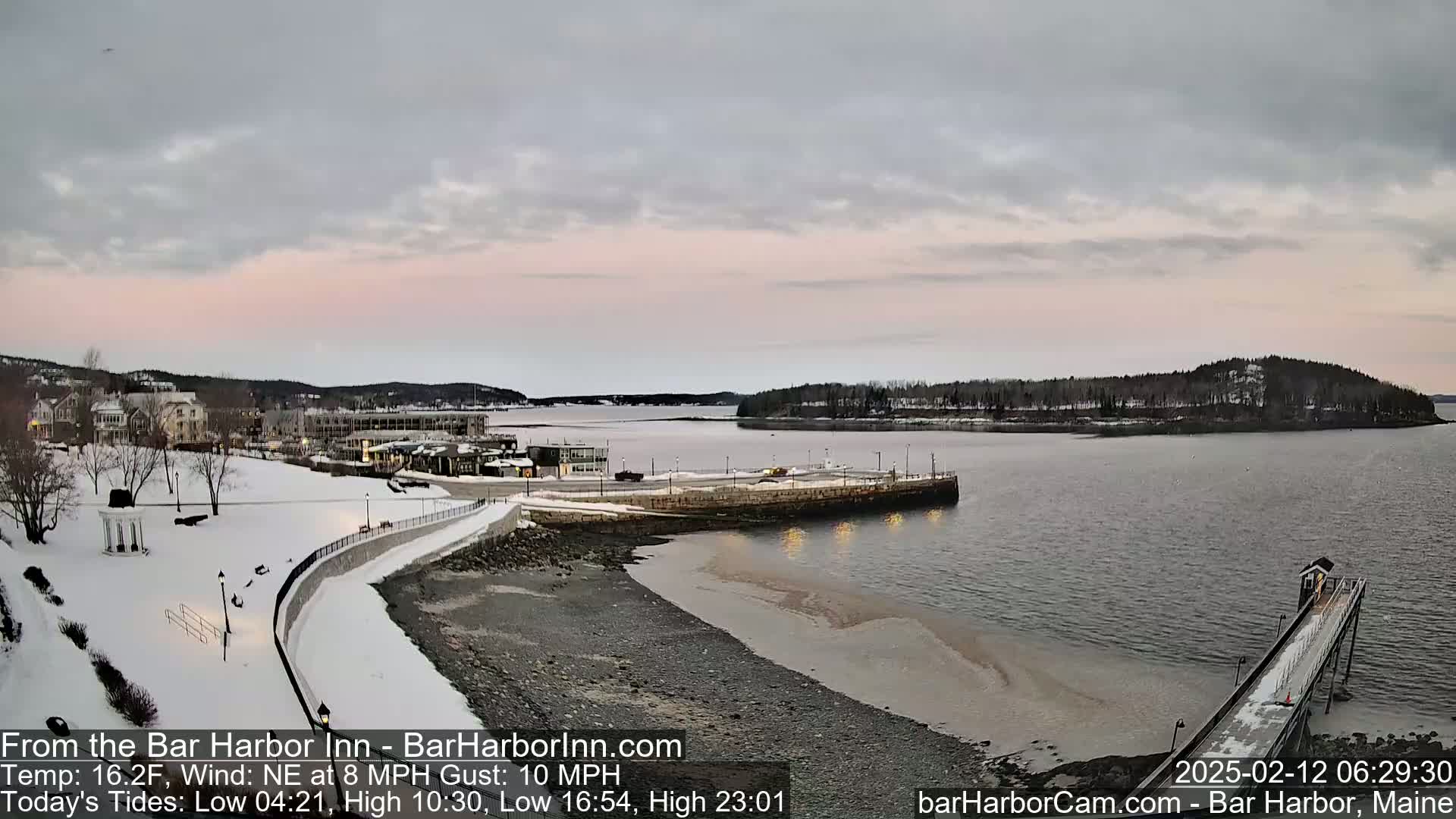 A snow-covered waterfront town with a calm bay under a pale pink and gray sky.
