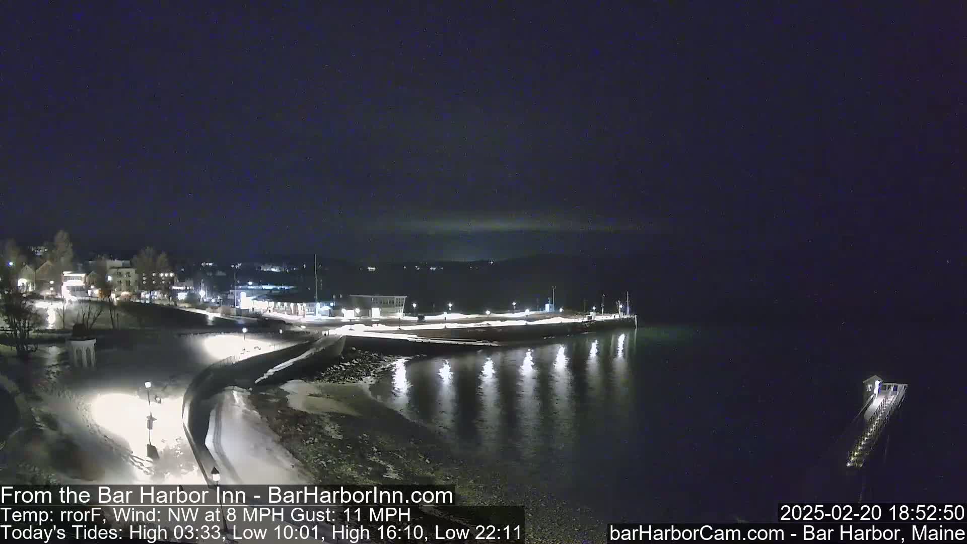 A nighttime view of a snow-covered waterfront town, with calm water reflecting lights and a pier extending into the dark bay under a clear sky.