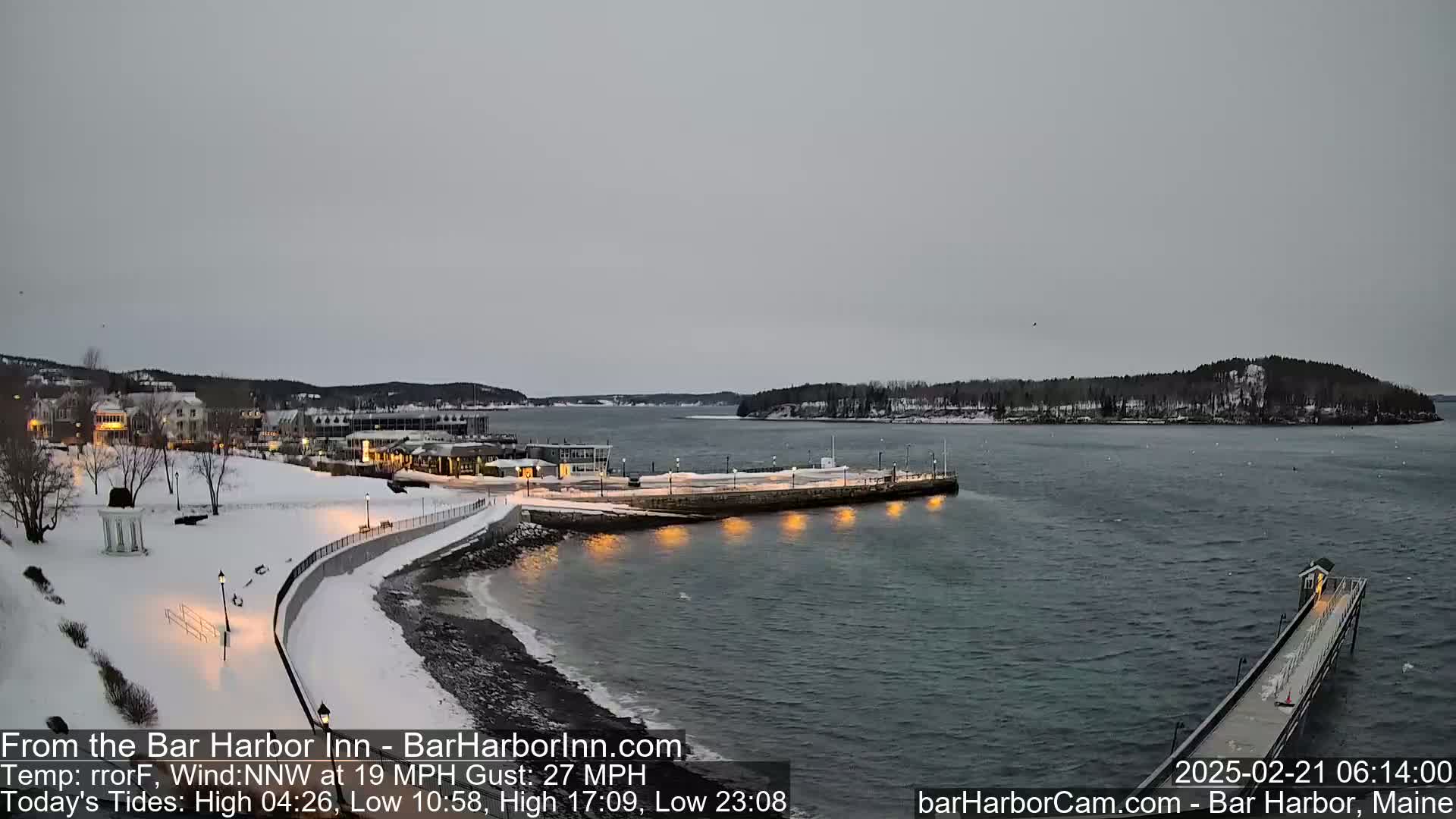 A snow-covered waterfront town is shown under an overcast sky, with a calm body of water, a long pier extending into the water, and lights illuminating the shoreline at night.