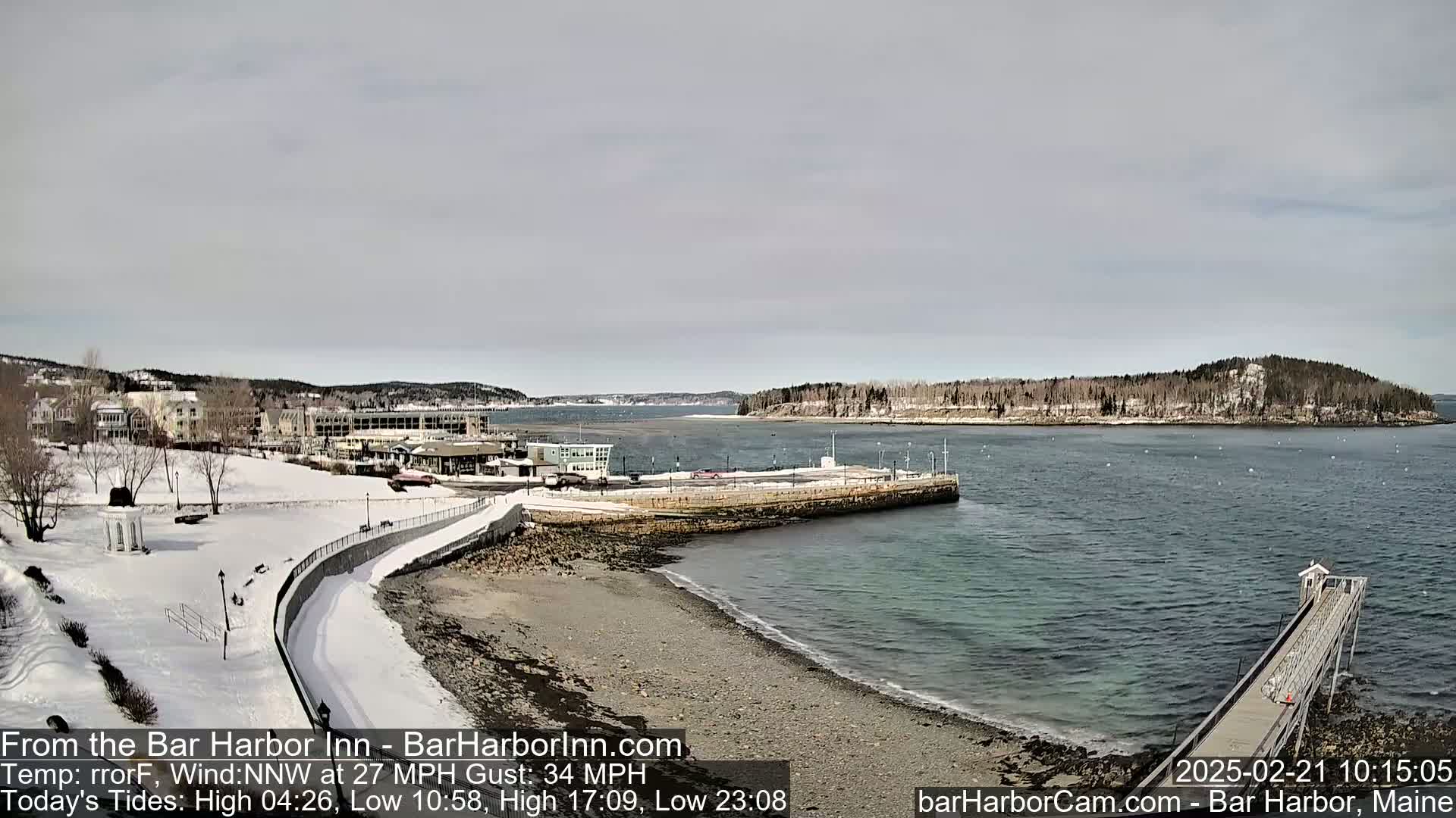 A snow-covered waterfront scene shows a harbor with buildings, a partially frozen shoreline, and a long pier extending into calm water under a cloudy sky.