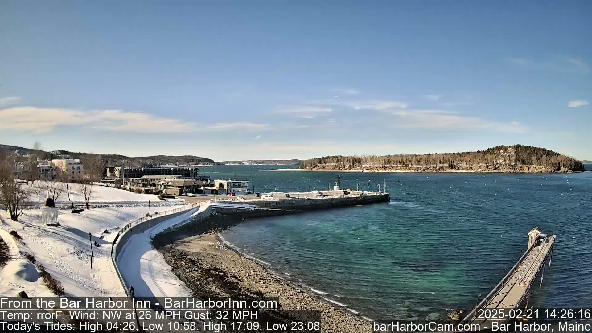 A snow-covered shoreline curves around a harbor with a long pier extending into clear, teal water under a mostly sunny sky.