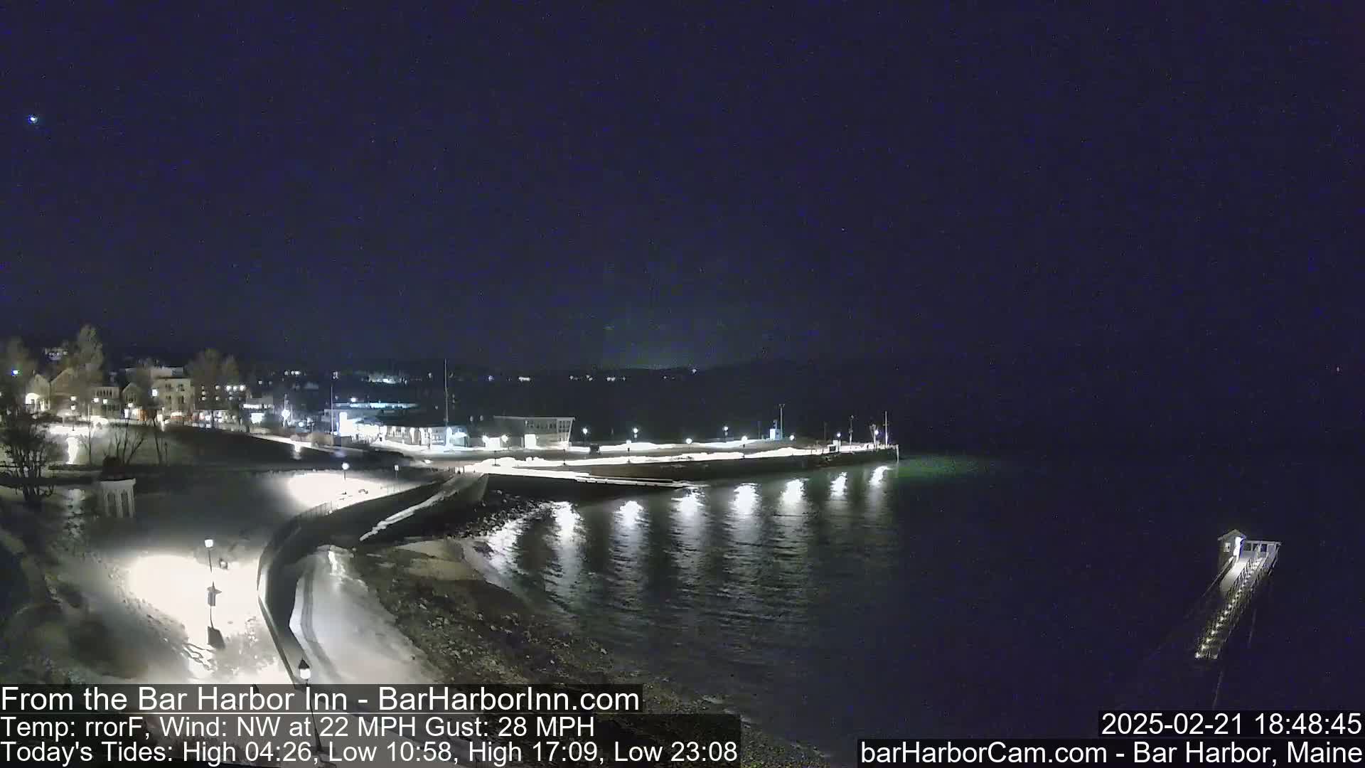 A nighttime, snow-covered waterfront scene shows a town illuminated under a dark sky with a 22 mph northwest wind and gusts up to 28 mph.