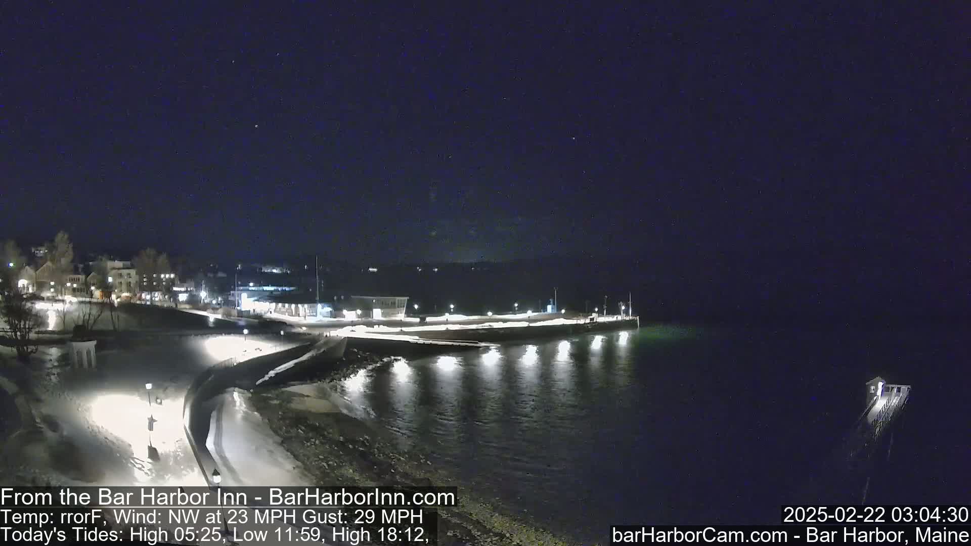 A nighttime view of a snow-covered harbor town with calm water under a clear, dark sky, with a northwest wind at 23 mph and gusts up to 29 mph.
