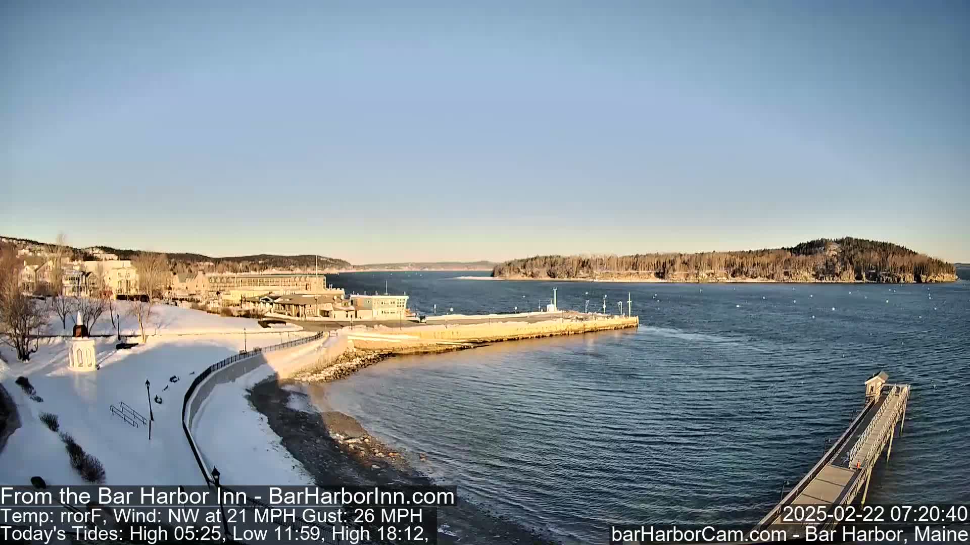 A snow-covered waterfront scene shows buildings, a harbor with a pier extending into the water, and a clear, sunny sky under a northwest wind.
