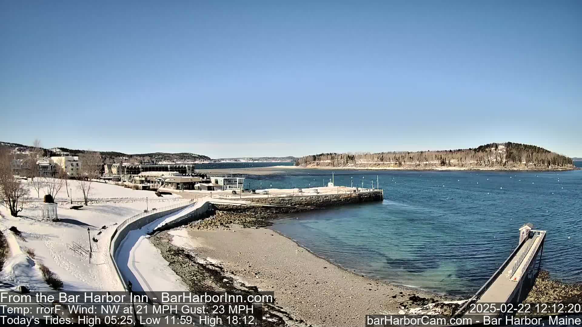 A snow-covered shoreline with a partially frozen body of water, featuring buildings and a long pier under a clear blue sky.