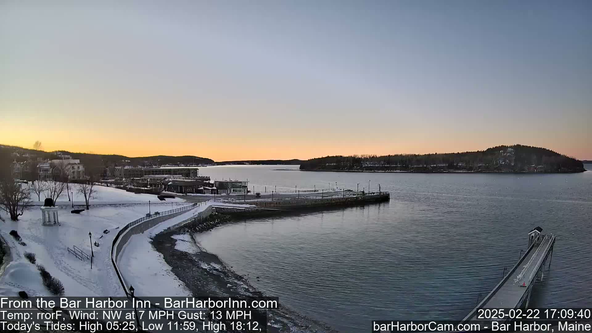 A snow-covered waterfront town is seen at sunset, with a calm body of water and a long pier extending into it.