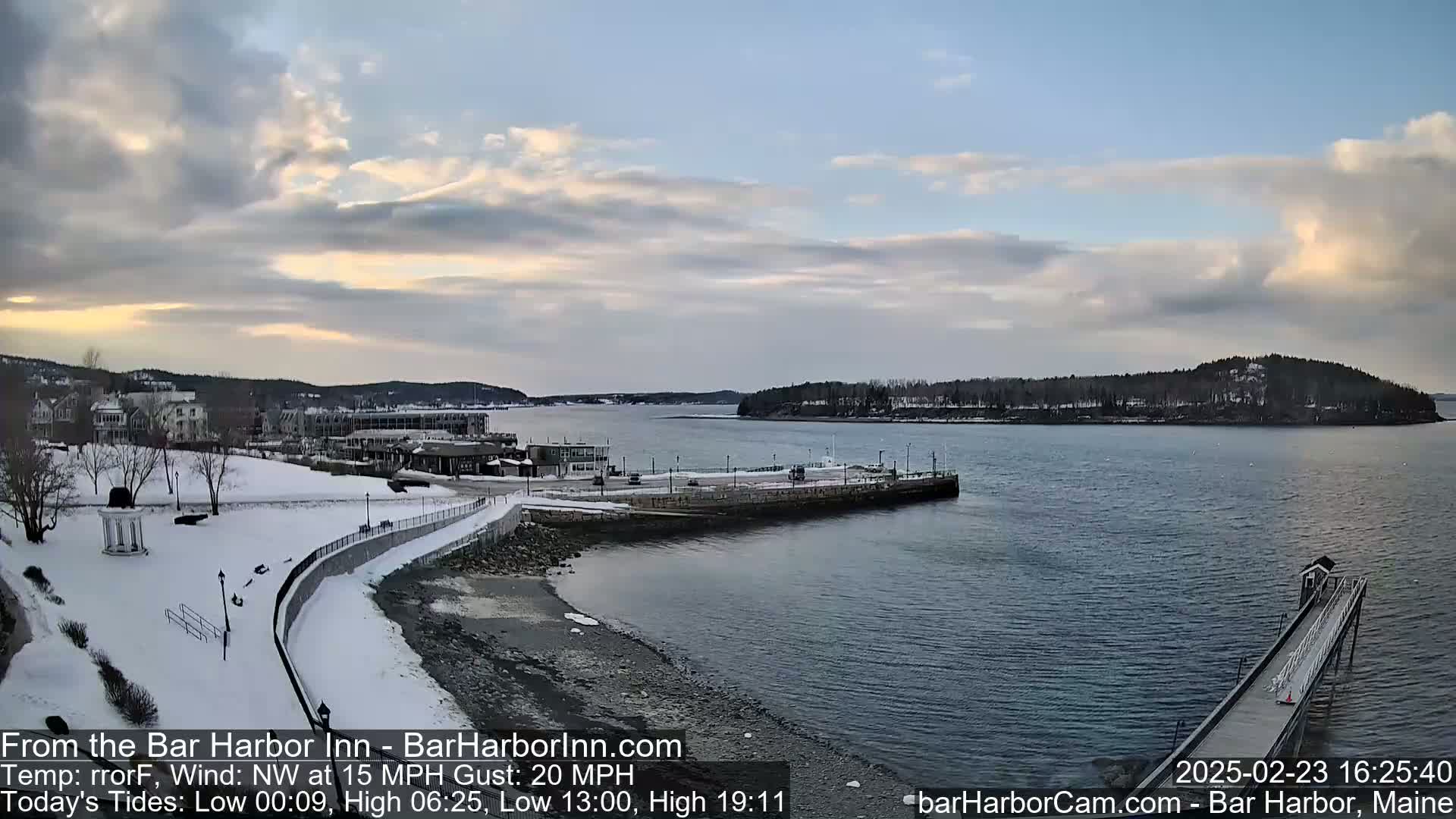 A snow-covered waterfront scene shows a calm body of water with a long pier extending into it, buildings in the background under a partly cloudy sky.