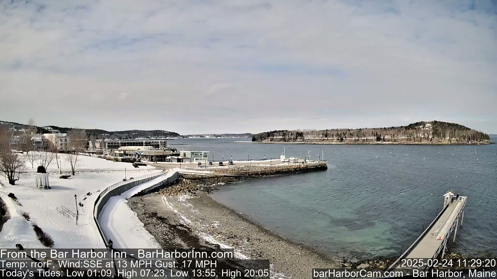 A snow-covered waterfront scene shows a harbor with a long pier extending into the water, buildings along the shore, and a small island in the distance under a partly cloudy sky.