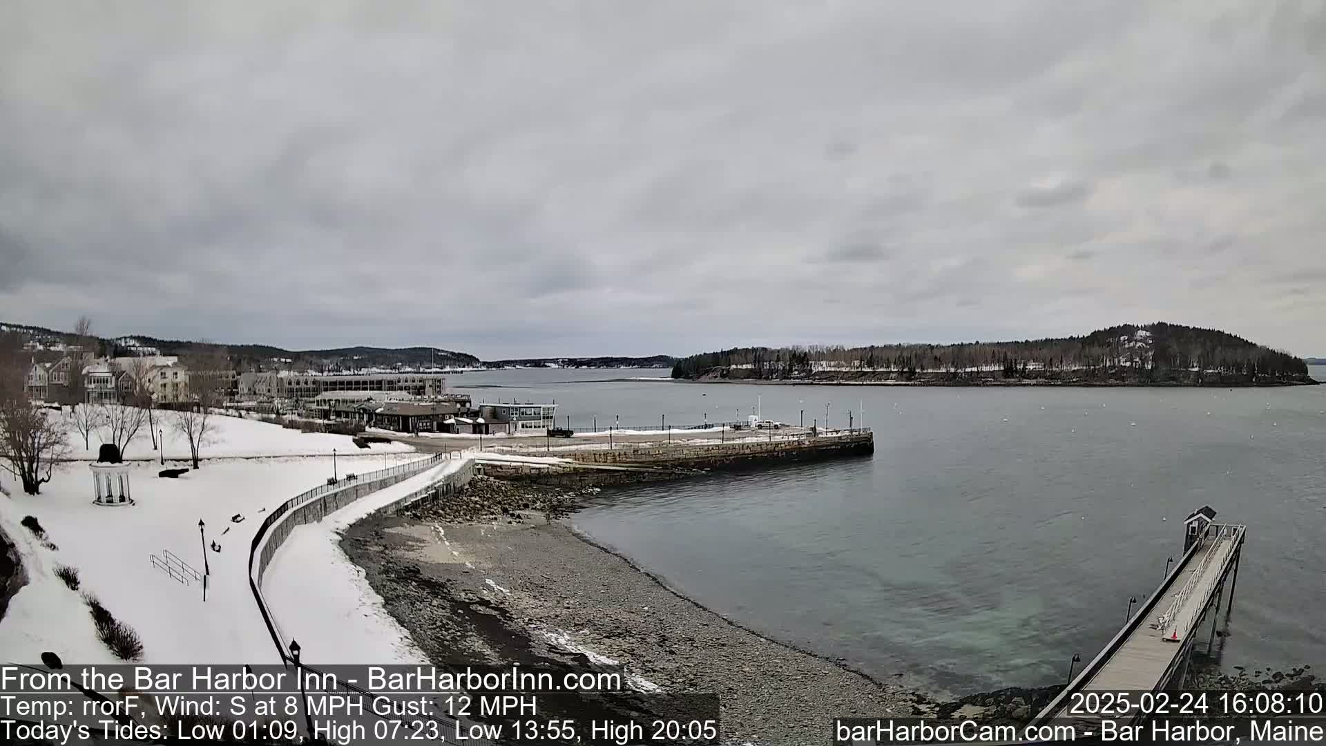A snow-covered waterfront scene shows a town with buildings, a partly frozen harbor, and a long pier extending into the calm, gray water under an overcast sky.