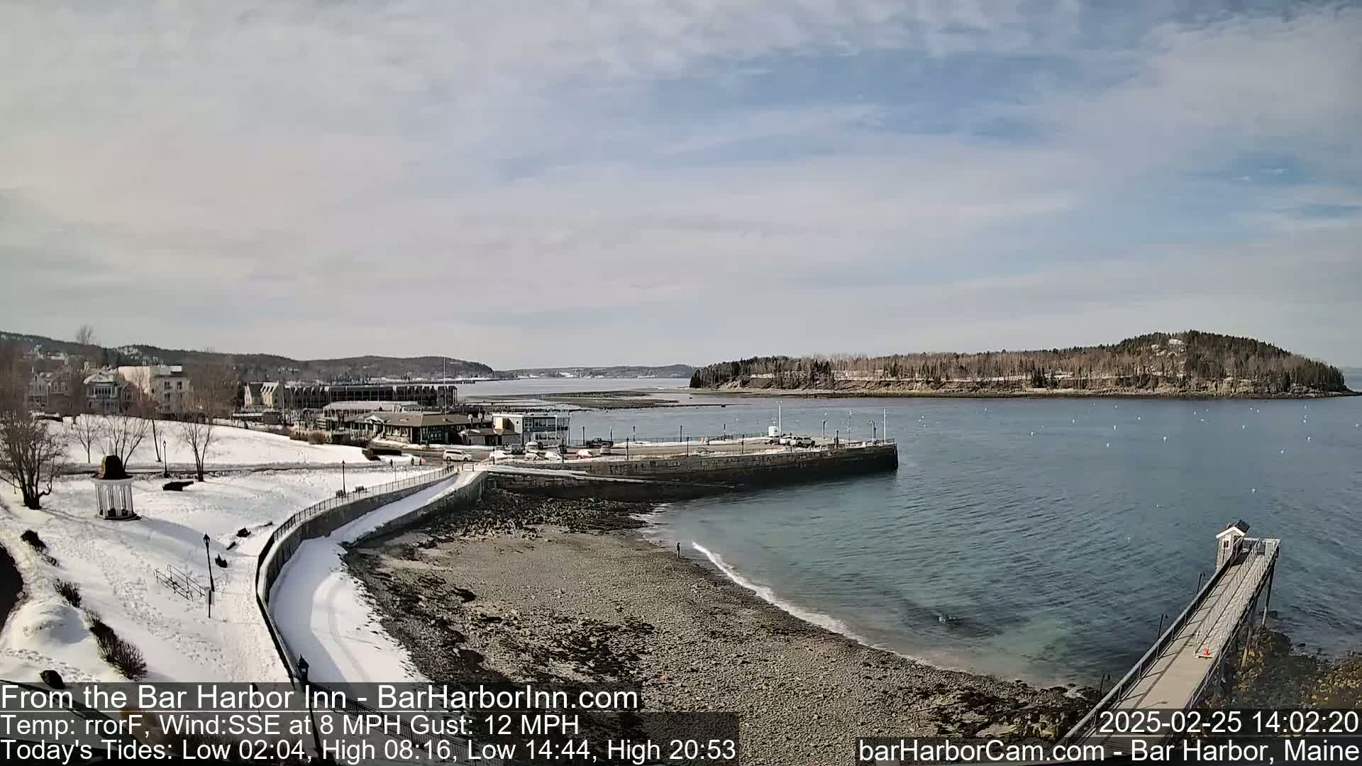 A snow-dusted coastal town is seen from a high vantage point on a partly cloudy day, overlooking a harbor with a small island and a long pier extending into the water.