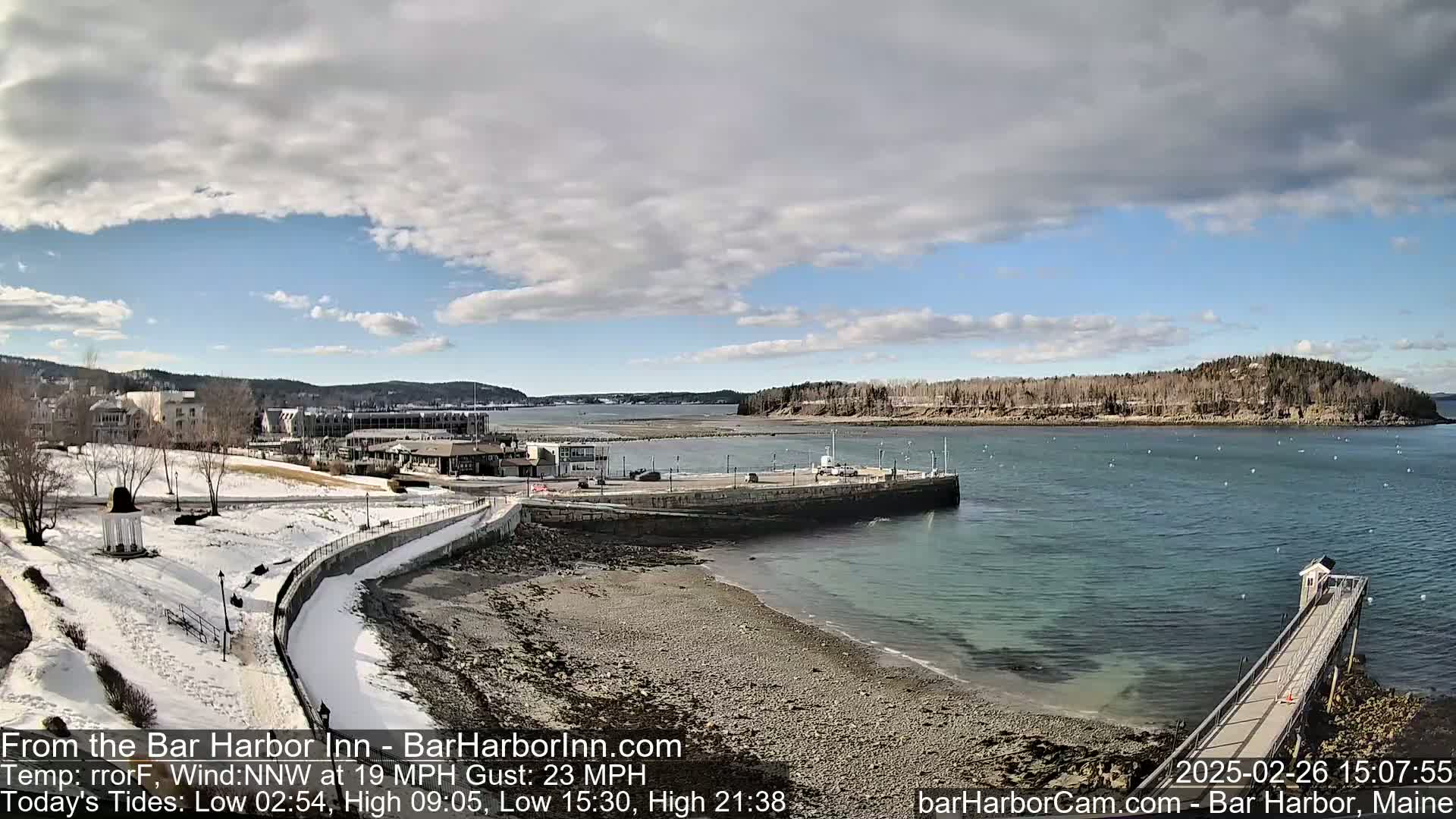 A snow-dusted waterfront scene shows a partially frozen harbor with a small pier extending into the calm water under a partly cloudy sky.