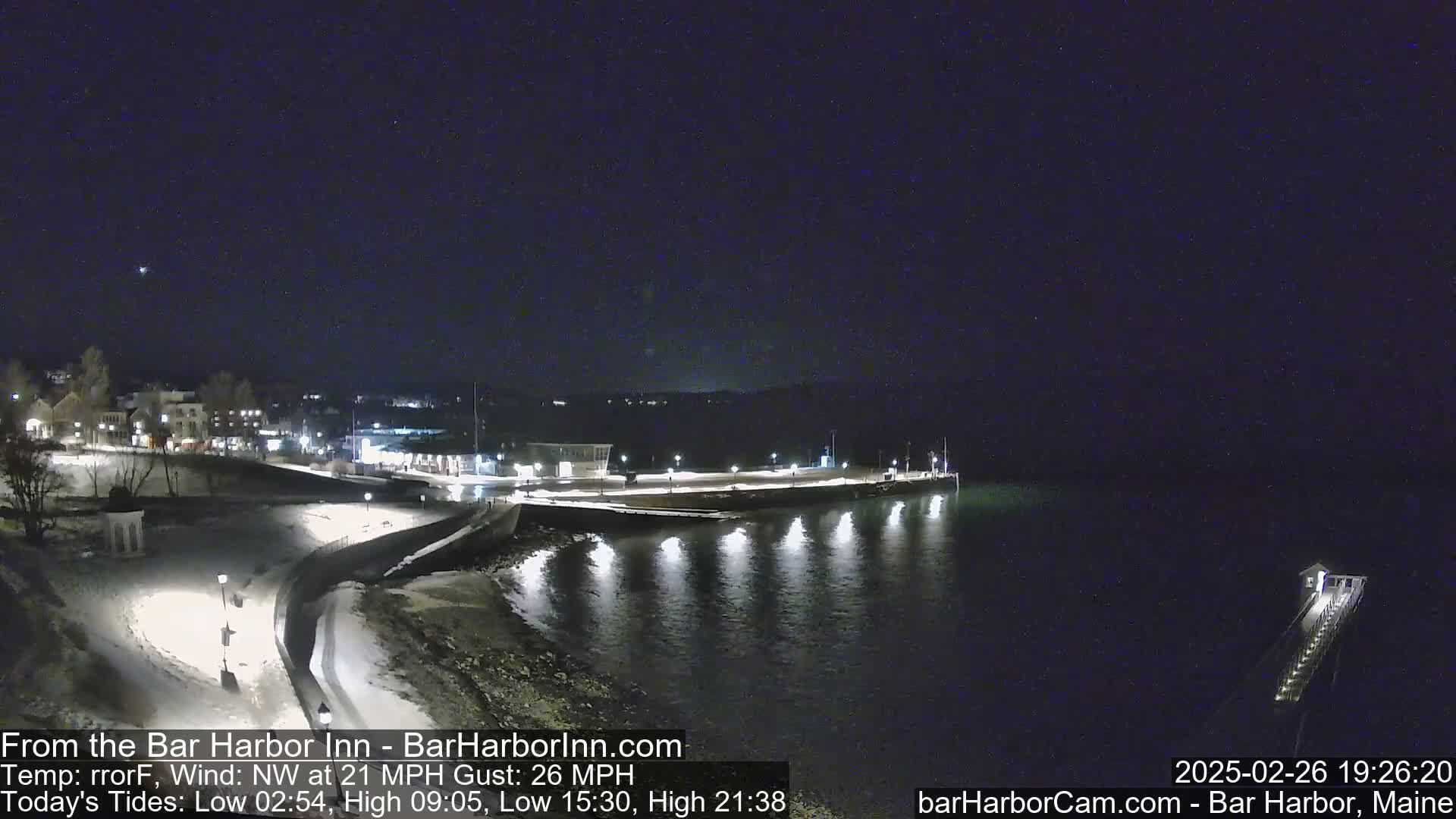 A nighttime view of a snow-dusted waterfront, with a pier extending into dark water under a clear sky, and a town visible in the background under a northwest wind of 21 mph with gusts up to 26 mph.
