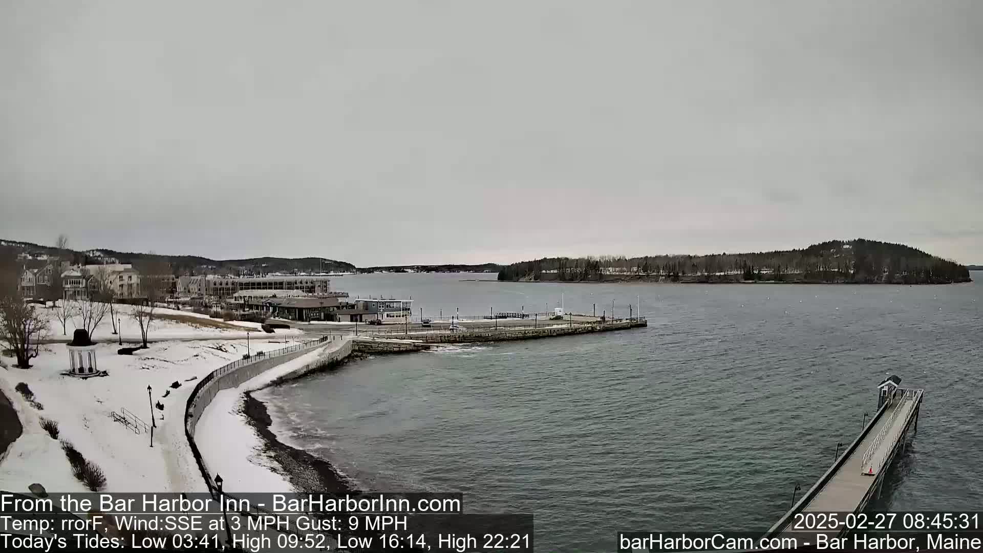 A snow-covered waterfront town with a calm harbor is seen under an overcast sky.