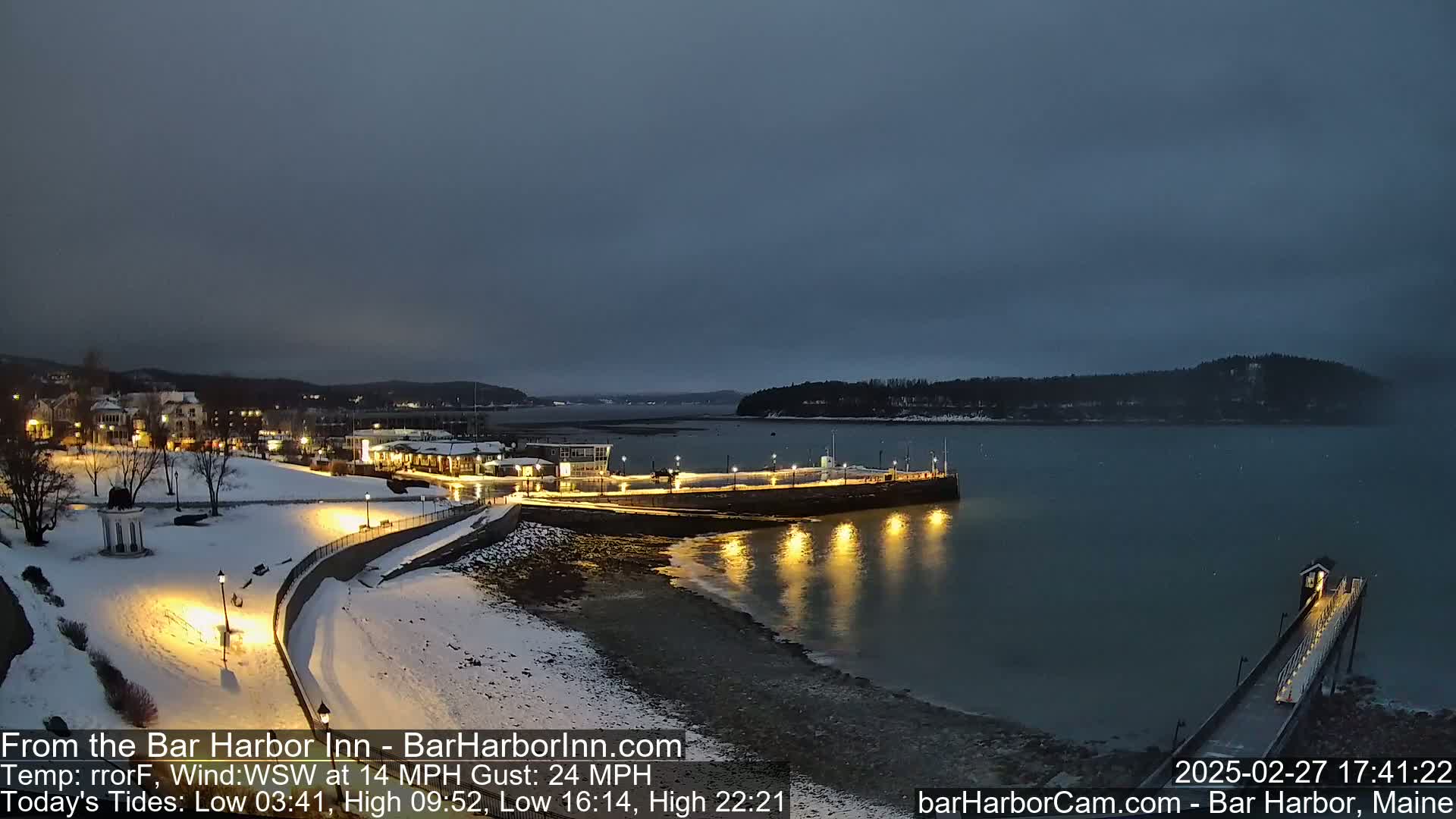 A snow-covered waterfront town is illuminated at dusk, with a calm body of water reflecting the lights.