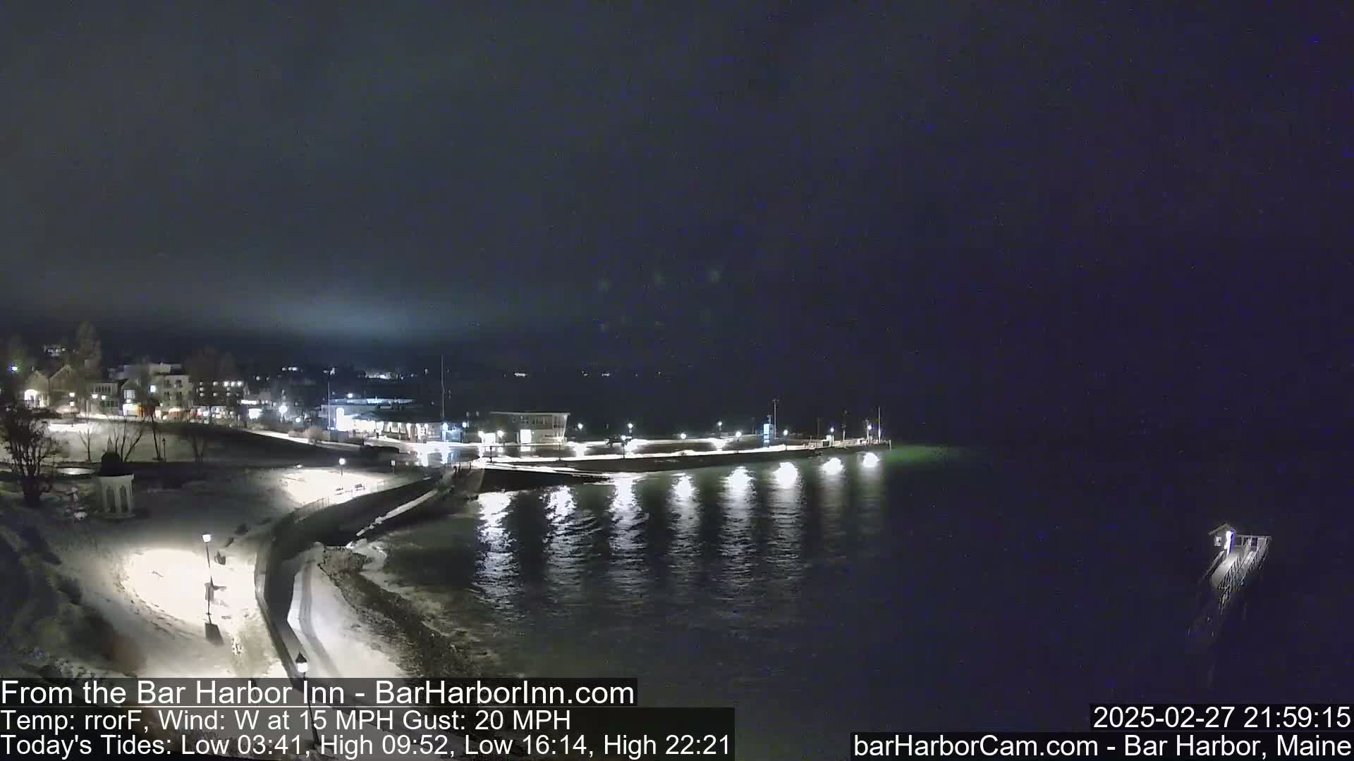 A snow-covered waterfront town is seen at night under a dark sky, with a calm body of water reflecting lights from a pier.