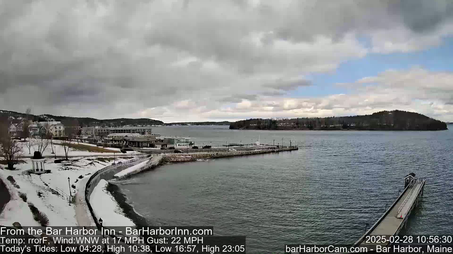 A partly cloudy day with a WNW wind at 17 mph and gusts up to 22 mph shows a snow-covered waterfront town with a harbor, small islands, and a long pier extending into the water.