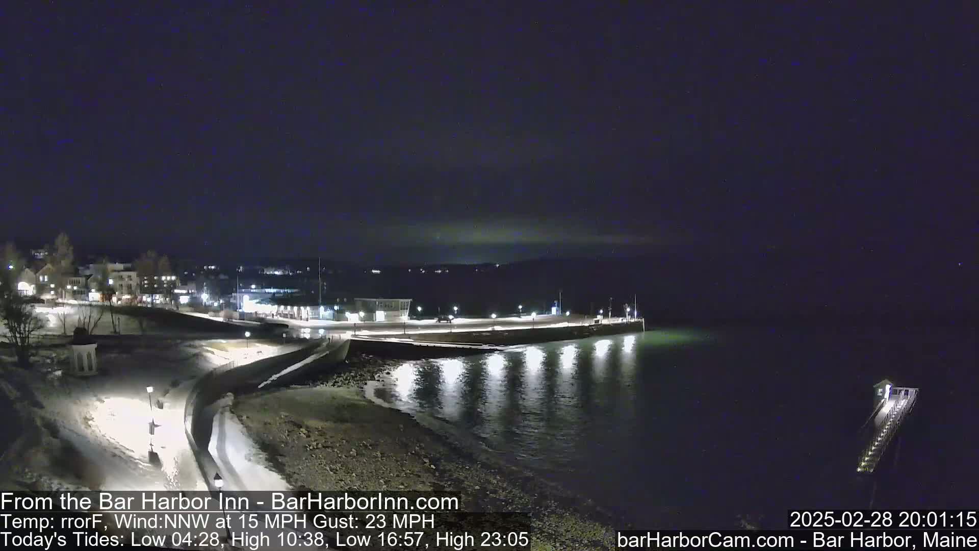 A nighttime view of a snow-dusted harbor town with a calm, dark body of water reflecting lights from the shoreline under a clear, dark sky.