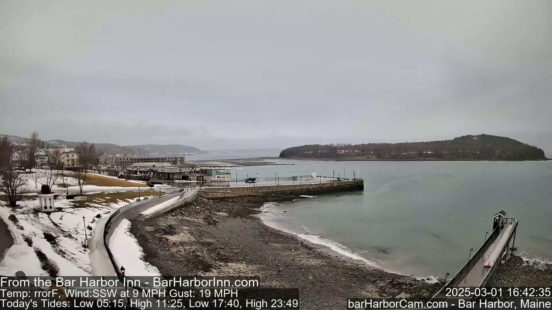A partly snowy coastal town is seen on an overcast day, featuring a rocky shoreline, a long pier extending into calm water, and an island in the distance.