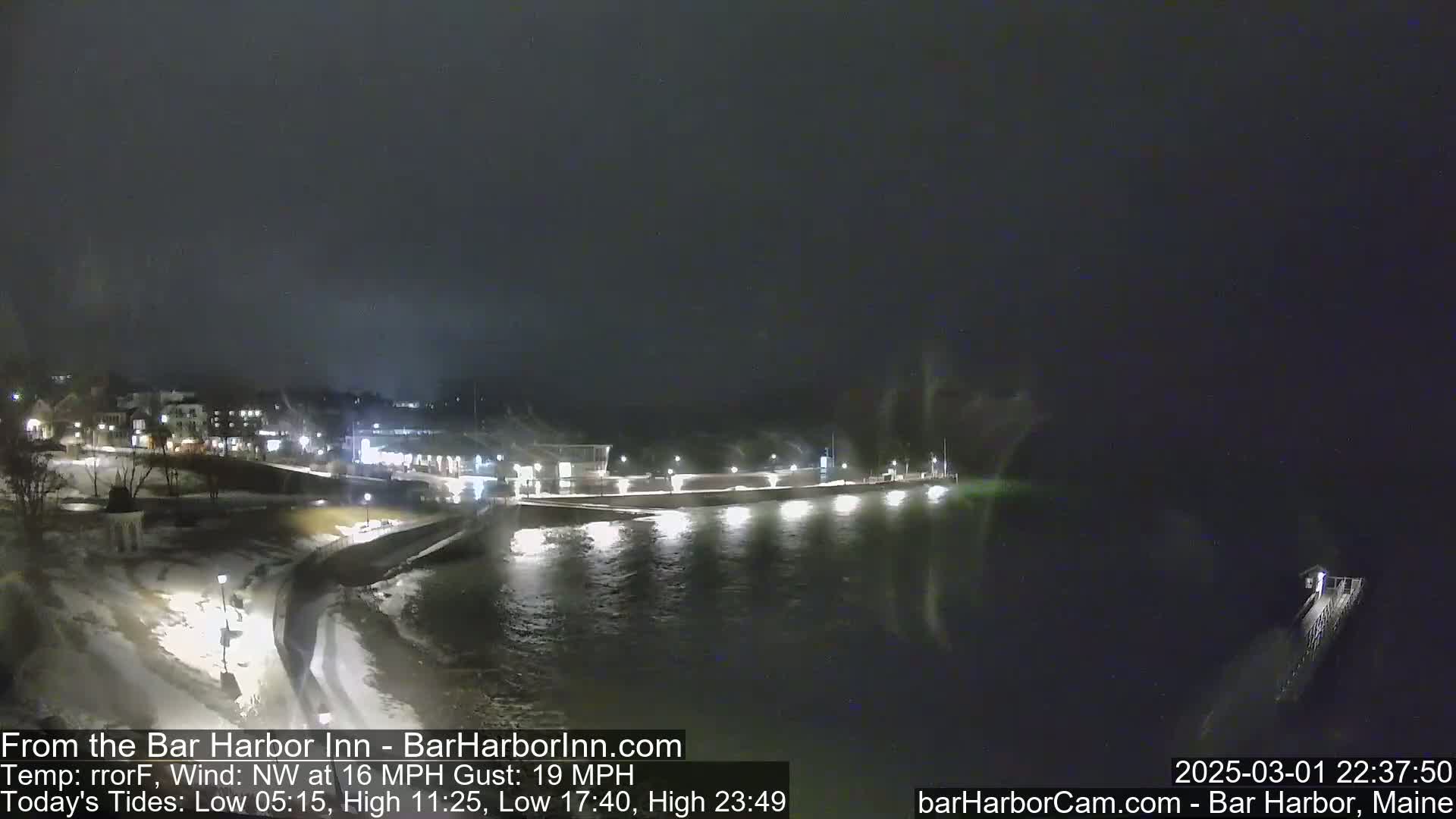 A nighttime view of a snow-covered waterfront town with a dark, choppy body of water under a dark sky.