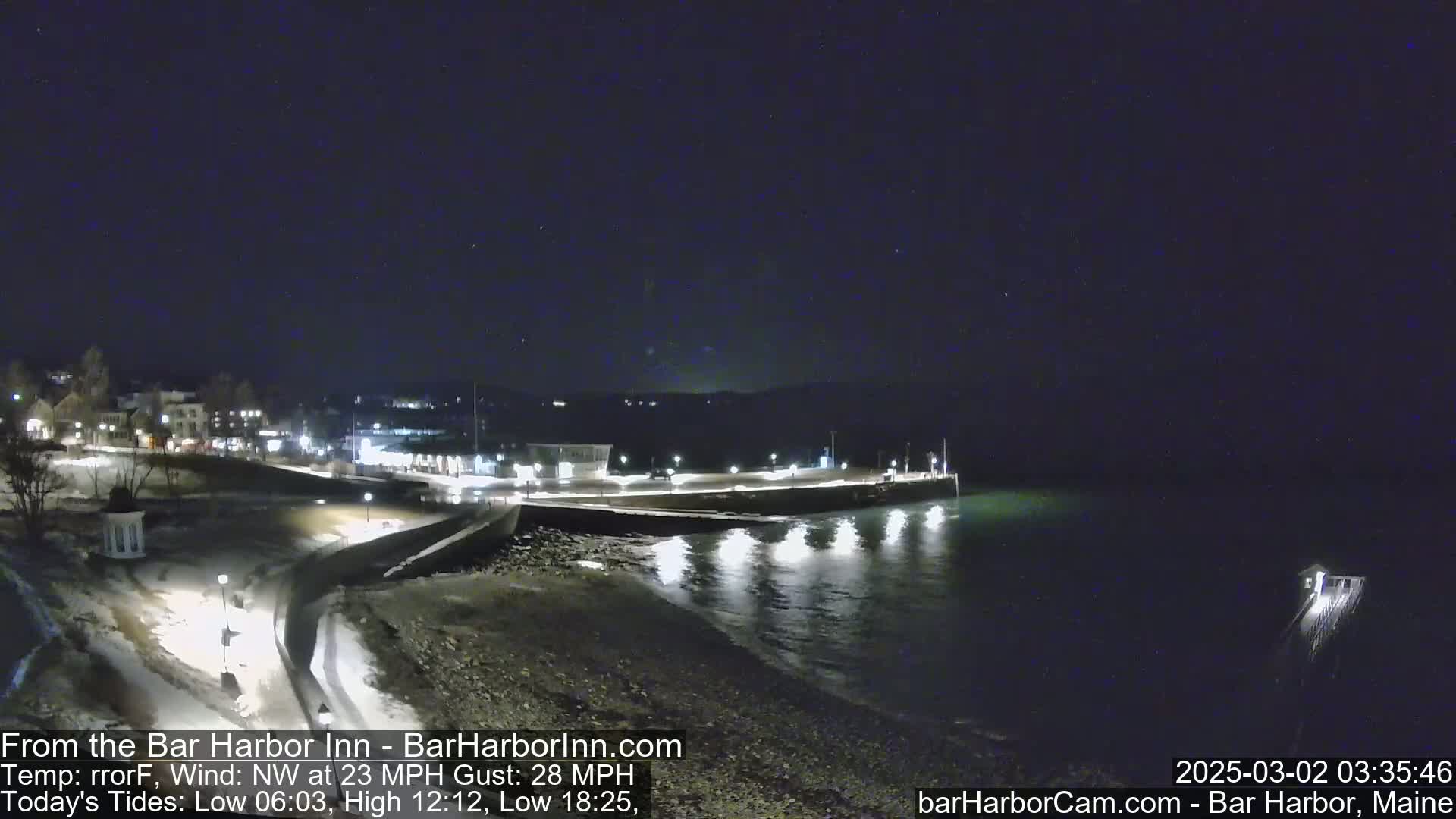 A nighttime, snow-dusted coastal town scene with a calm body of water under a dark, clear sky, featuring a pier extending into the water and buildings faintly illuminated along the shore.