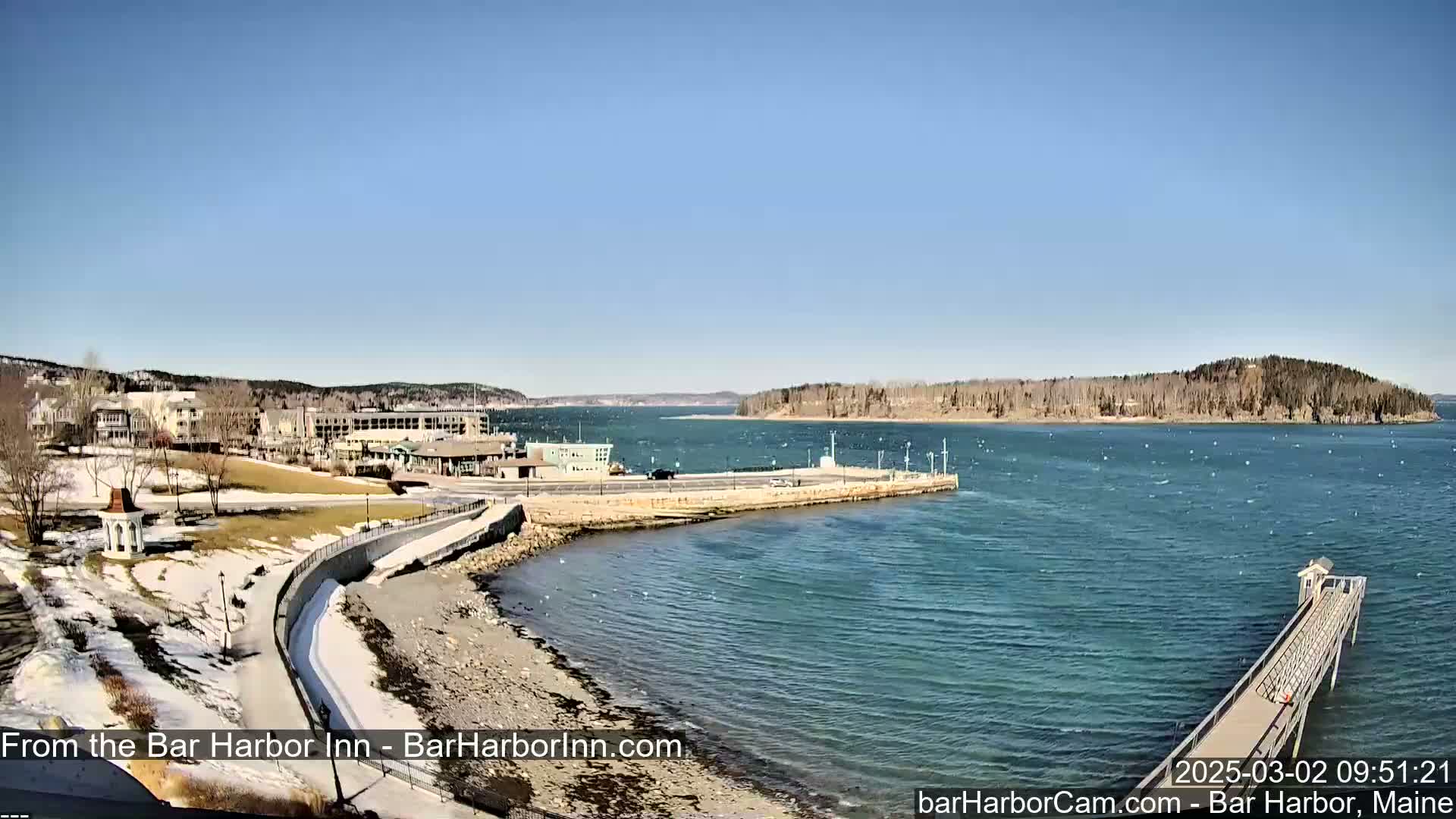 A clear, sunny day shows a coastal scene with a partially snow-covered shoreline, buildings, a long pier extending into the water, and a small island in the distance.