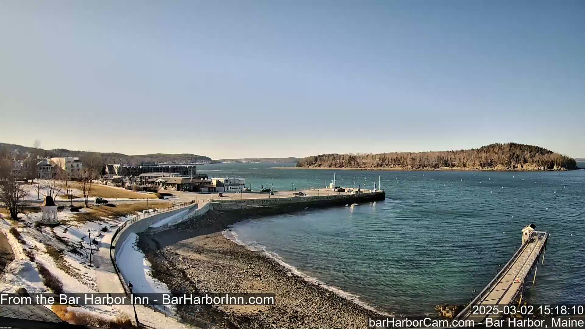 A waterfront scene on a sunny day shows a rocky shoreline, a parking area with a few vehicles, buildings, and a long pier extending into the calm water.