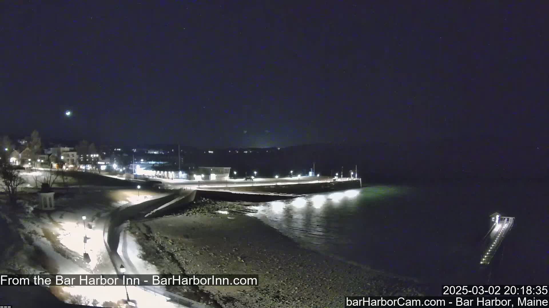 A nighttime coastal scene shows a snow-covered shoreline with buildings and lights in the distance, under a dark, clear sky.