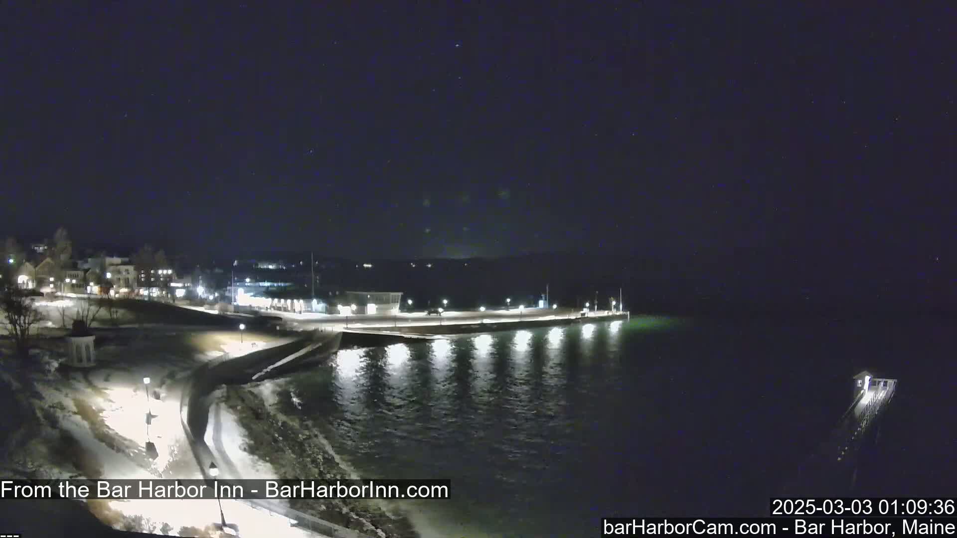 A nighttime view of a snow-covered waterfront, showing a town lit up across the water under a clear, starry sky.