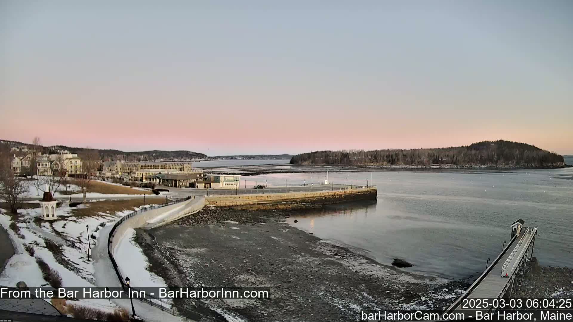 A waterfront view at dawn shows a harbor with buildings, a stone seawall, a pier, and a small island in the distance, under a pale pink sky.