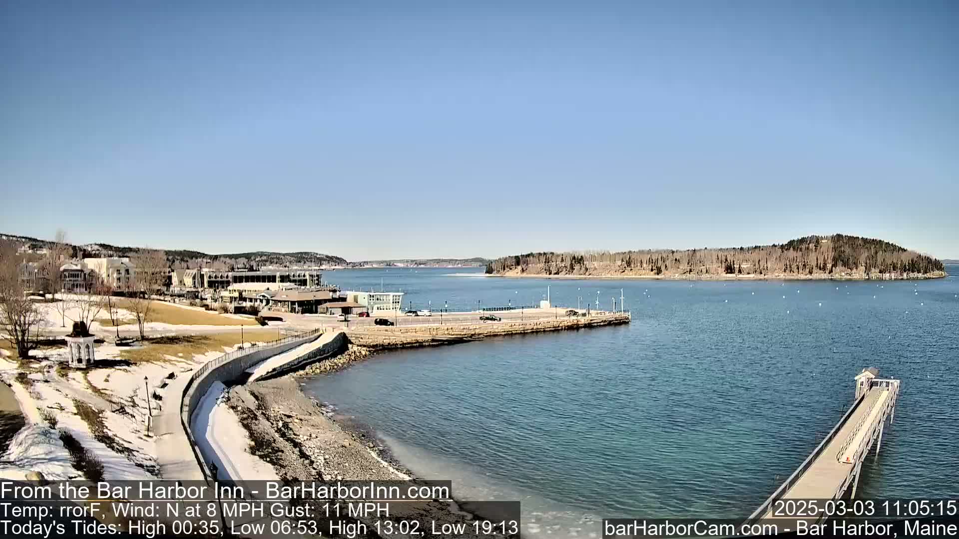 A waterfront view shows a harbor with a long pier extending into the water, buildings and a grassy area in the background, under a clear blue sky.