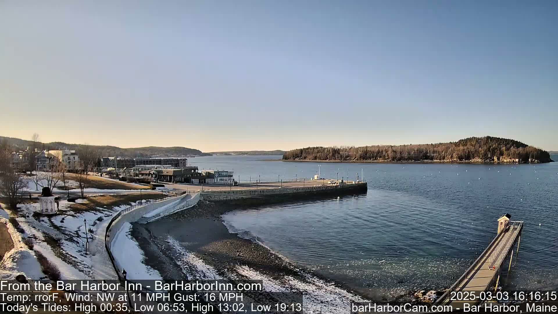 A coastal town scene on a partly sunny day with a northwest wind, featuring a harbor with a long pier extending into the water, a small island in the distance, and a shoreline with patches of snow.