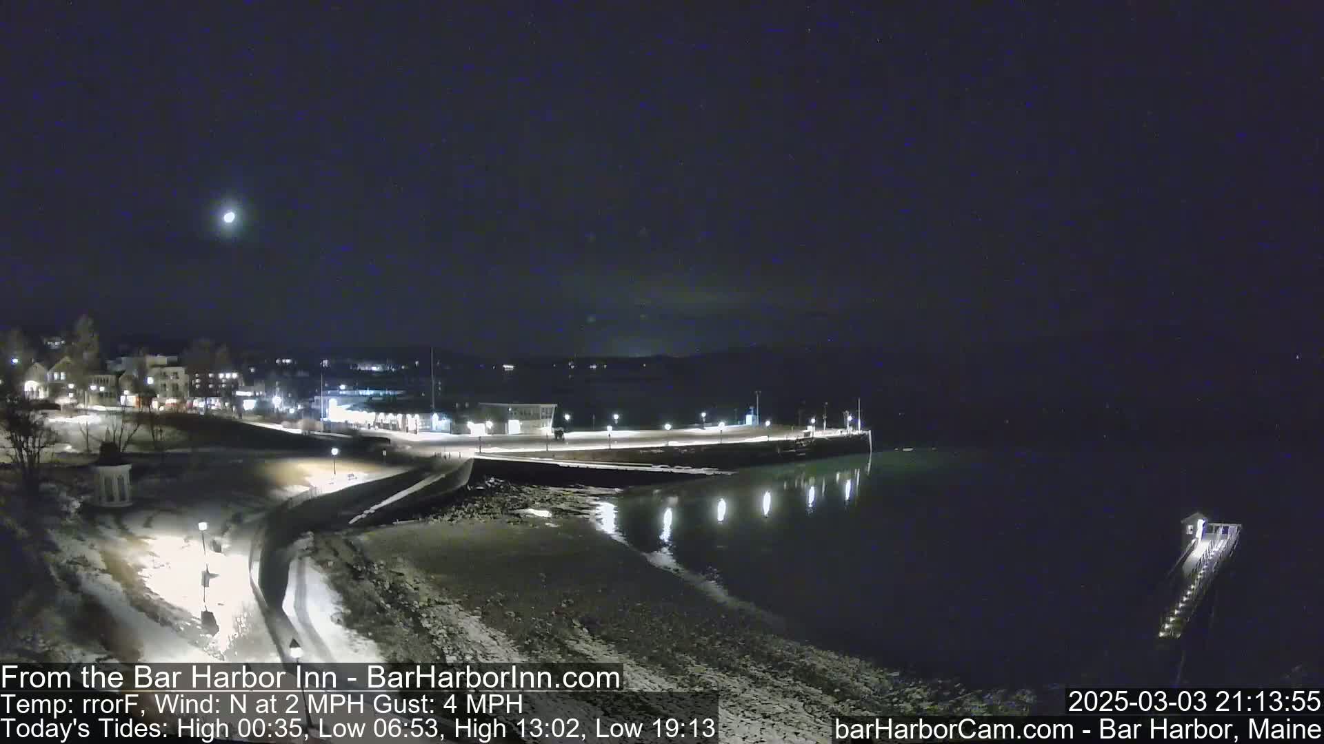 A nighttime view of a snow-covered waterfront town and harbor with calm water under a clear, moonlit sky.