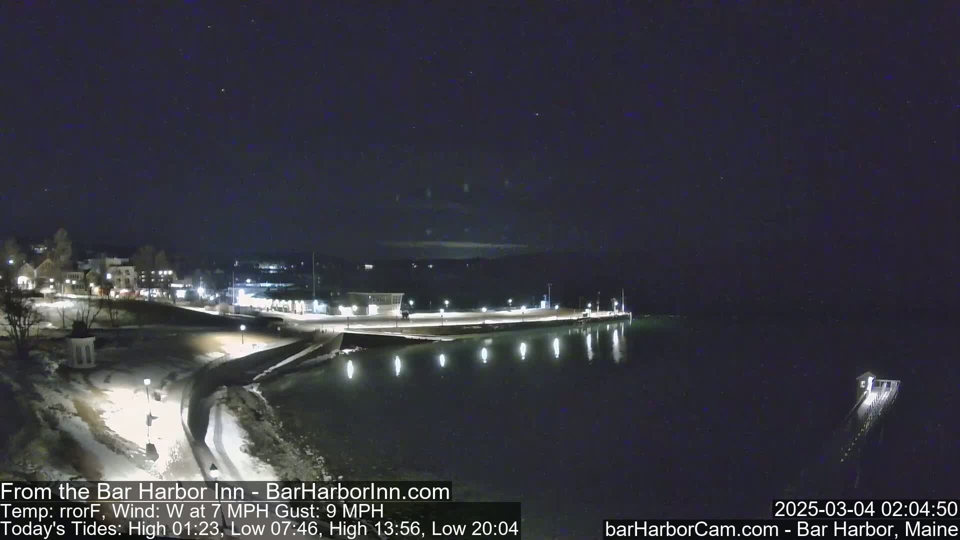 A nighttime, snow-dusted waterfront scene shows a calm body of water reflecting lights from a town and pier under a clear, dark sky.