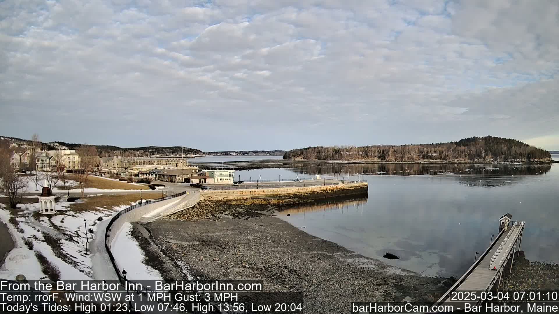 A partly cloudy day reveals a waterfront scene with a calm body of water, a stone walkway, buildings, and a small island in the distance; some snow is visible on the ground.