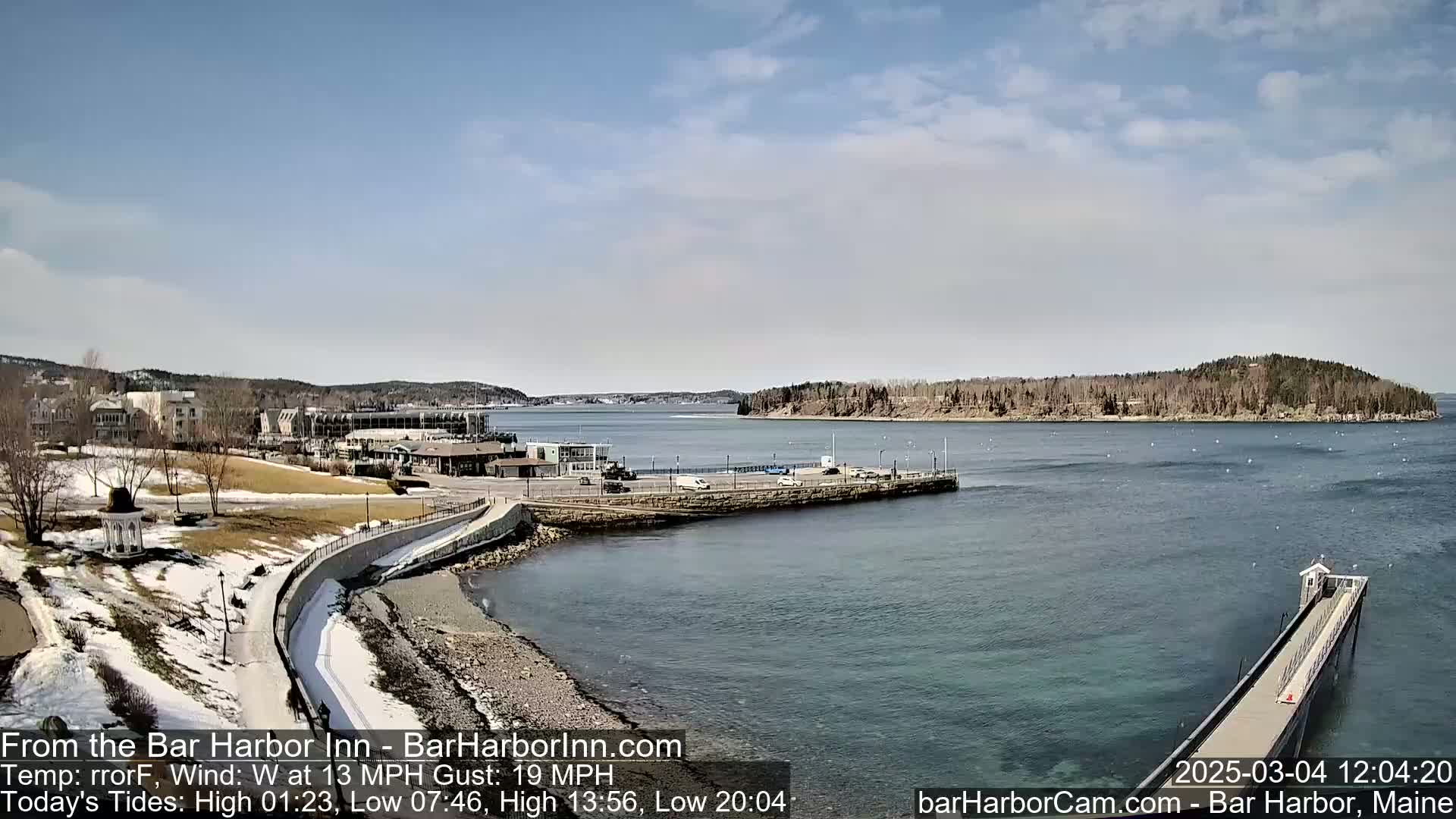 A partly sunny day shows a coastal town with a harbor, a long pier, and a small island in the distance, with some snow remaining on the ground near the water.