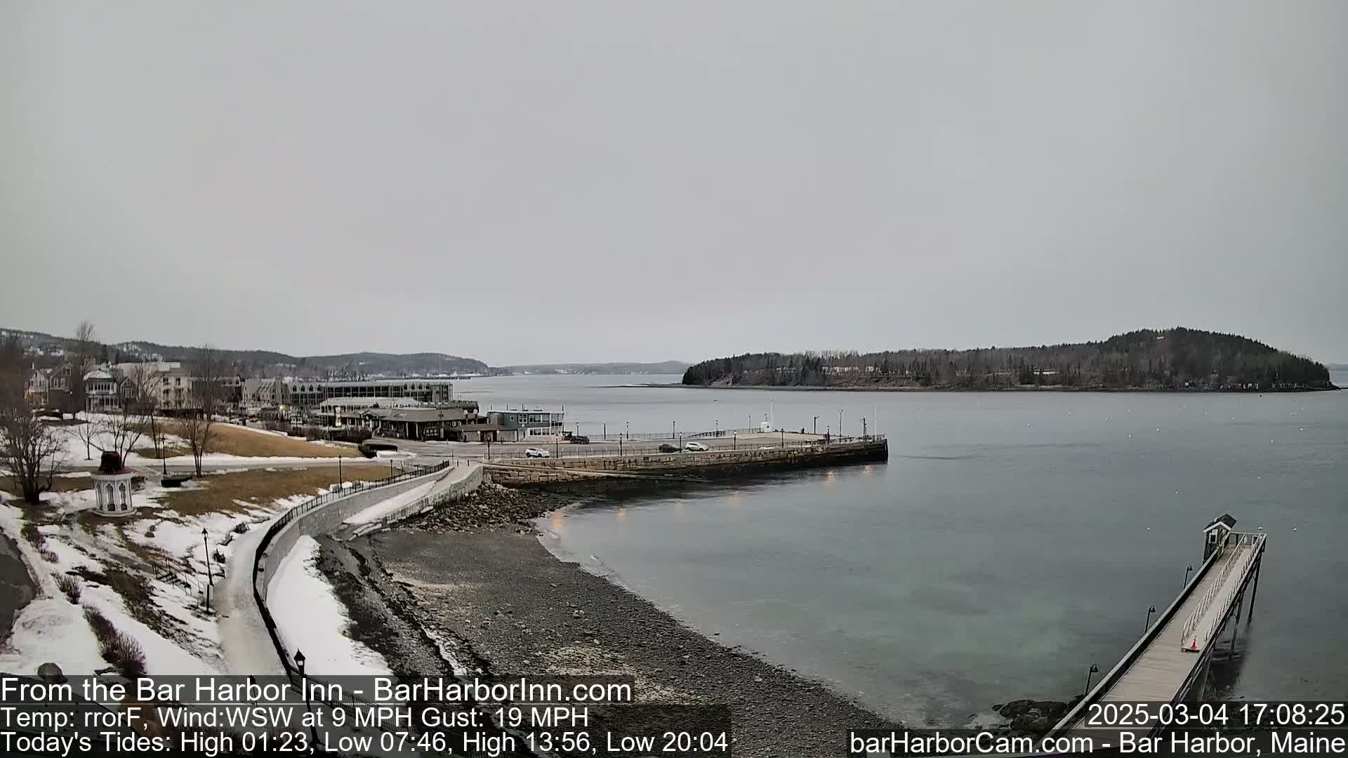 A partly-snowy waterfront scene shows a calm body of water with a long pier extending into it, under an overcast sky.