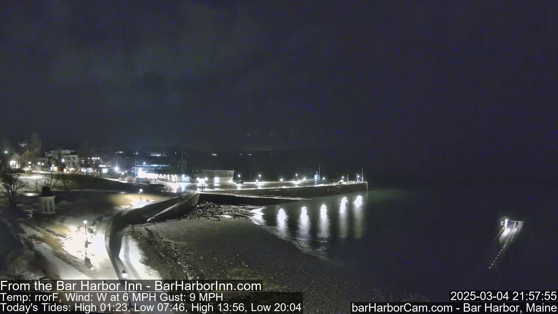 A nighttime view of a snow-covered shoreline with lights illuminating a town in the background under a dark, possibly slightly cloudy sky.