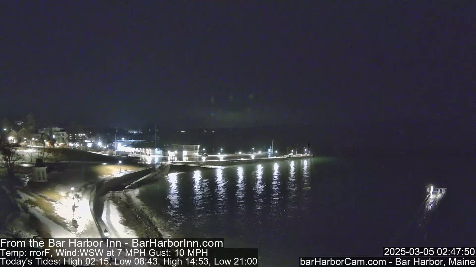 A nighttime view of a harbor with calm water reflecting lights from a waterfront building and pier under a dark, clear sky.