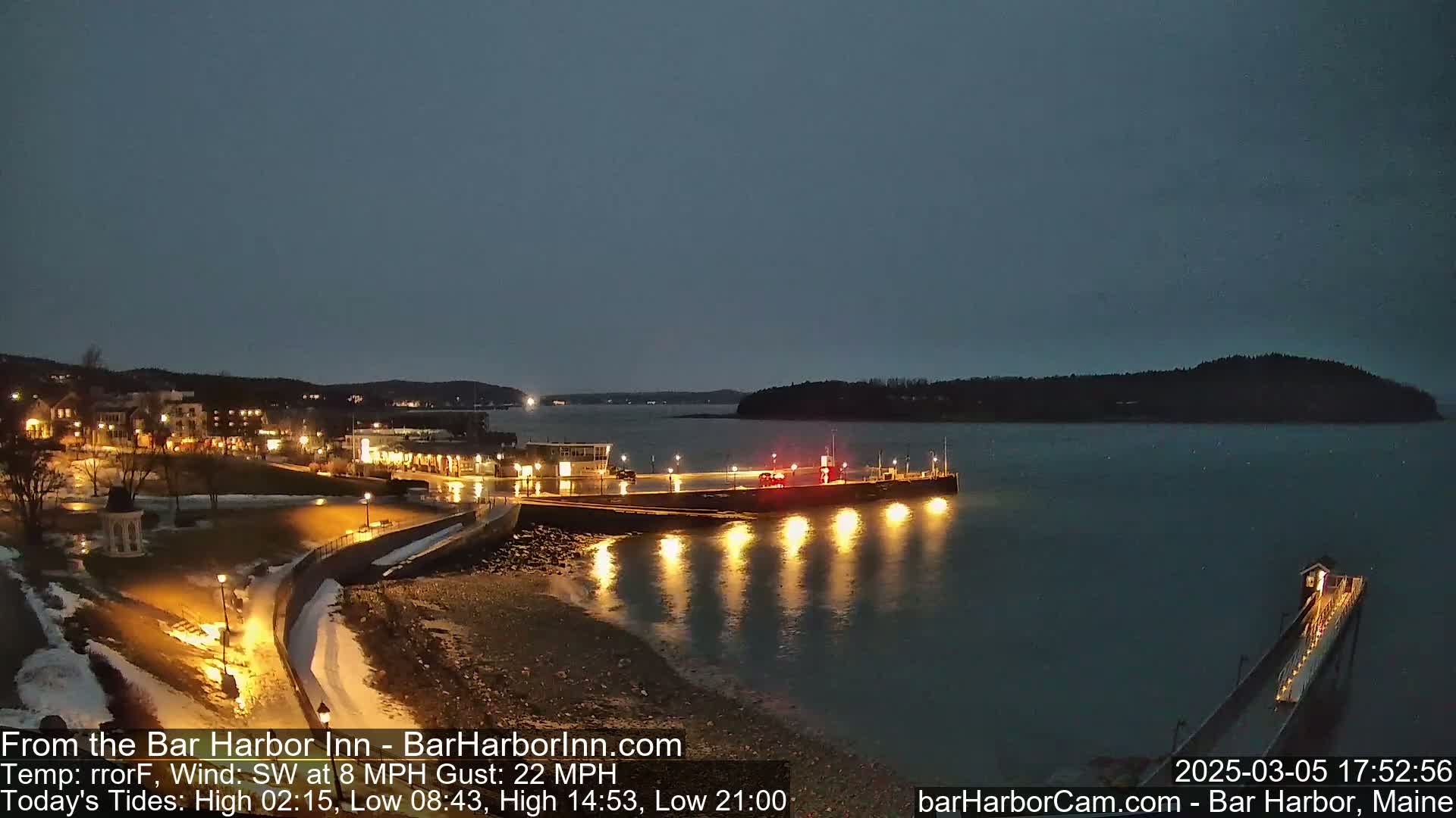 A nighttime view of a harbor town with illuminated buildings and piers, under a dark, mostly clear sky, with a slight southwest wind.