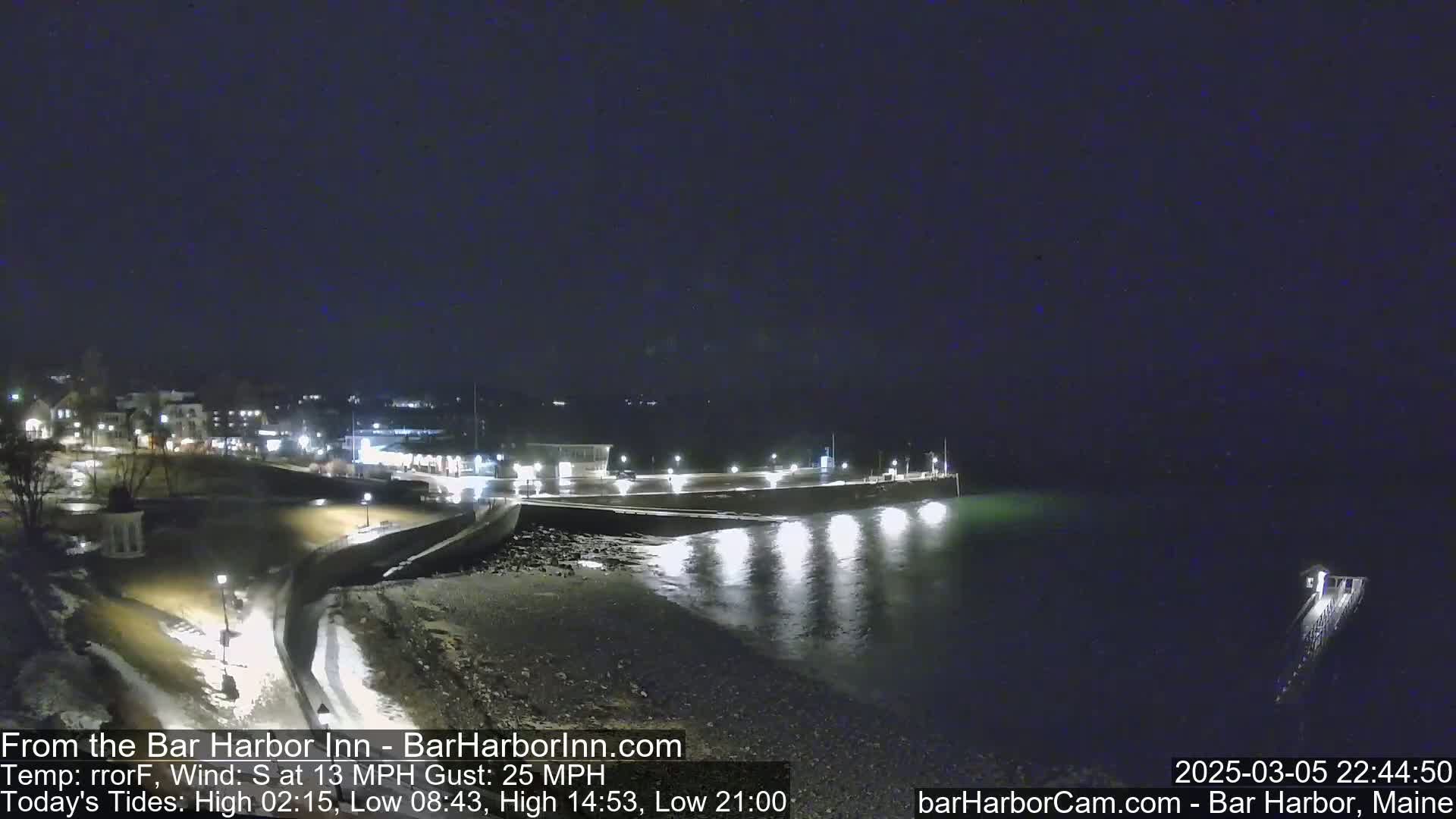 A nighttime coastal scene shows a dark, calm body of water reflecting lights from a nearby town and pier under a clear, dark sky.