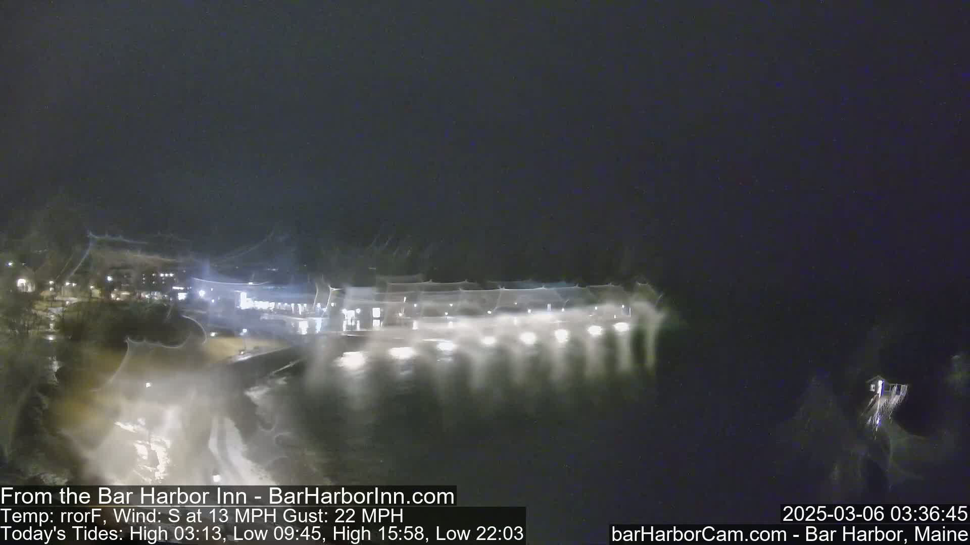A nighttime view of a waterfront area with buildings and lights, showing a dark, windy, and possibly rainy or wet scene.