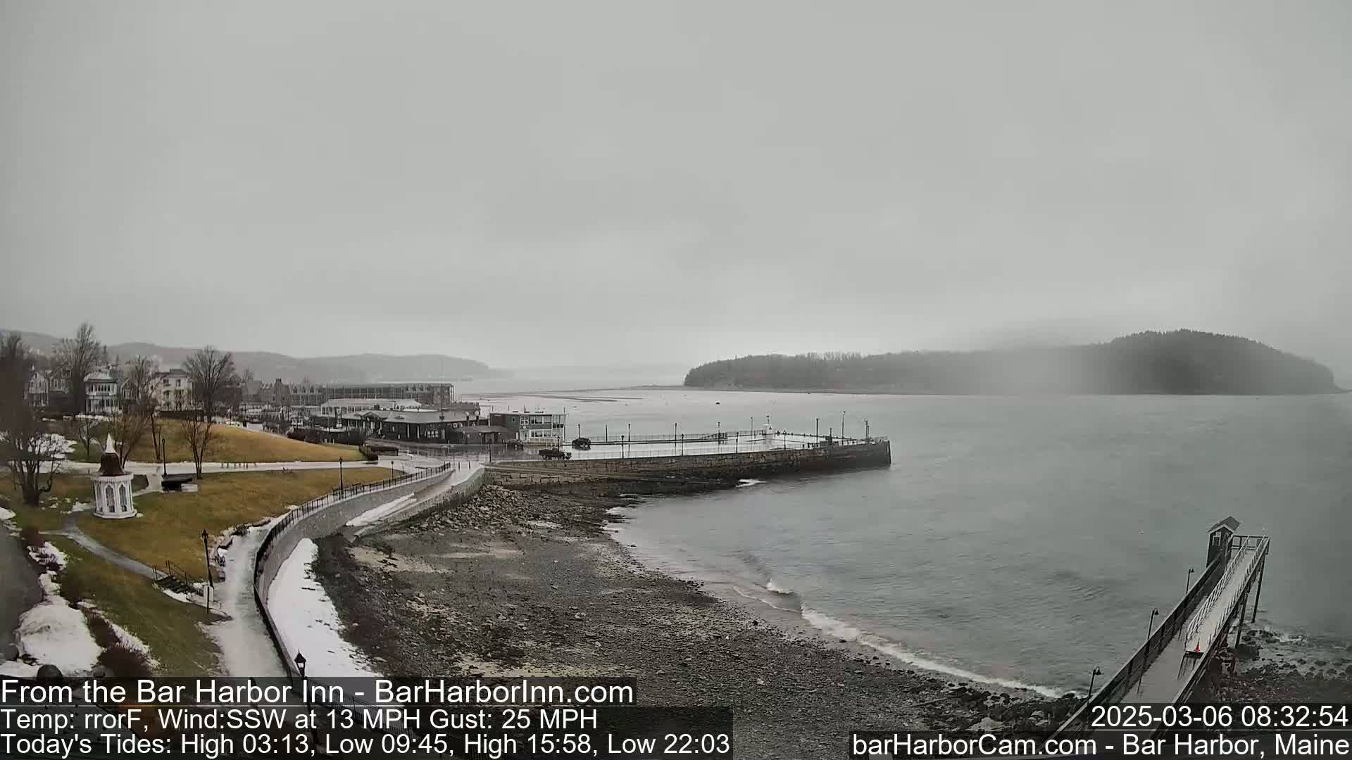 A coastal town, partially covered in snow, is seen under an overcast sky, with a calm body of water and a long pier extending into it.