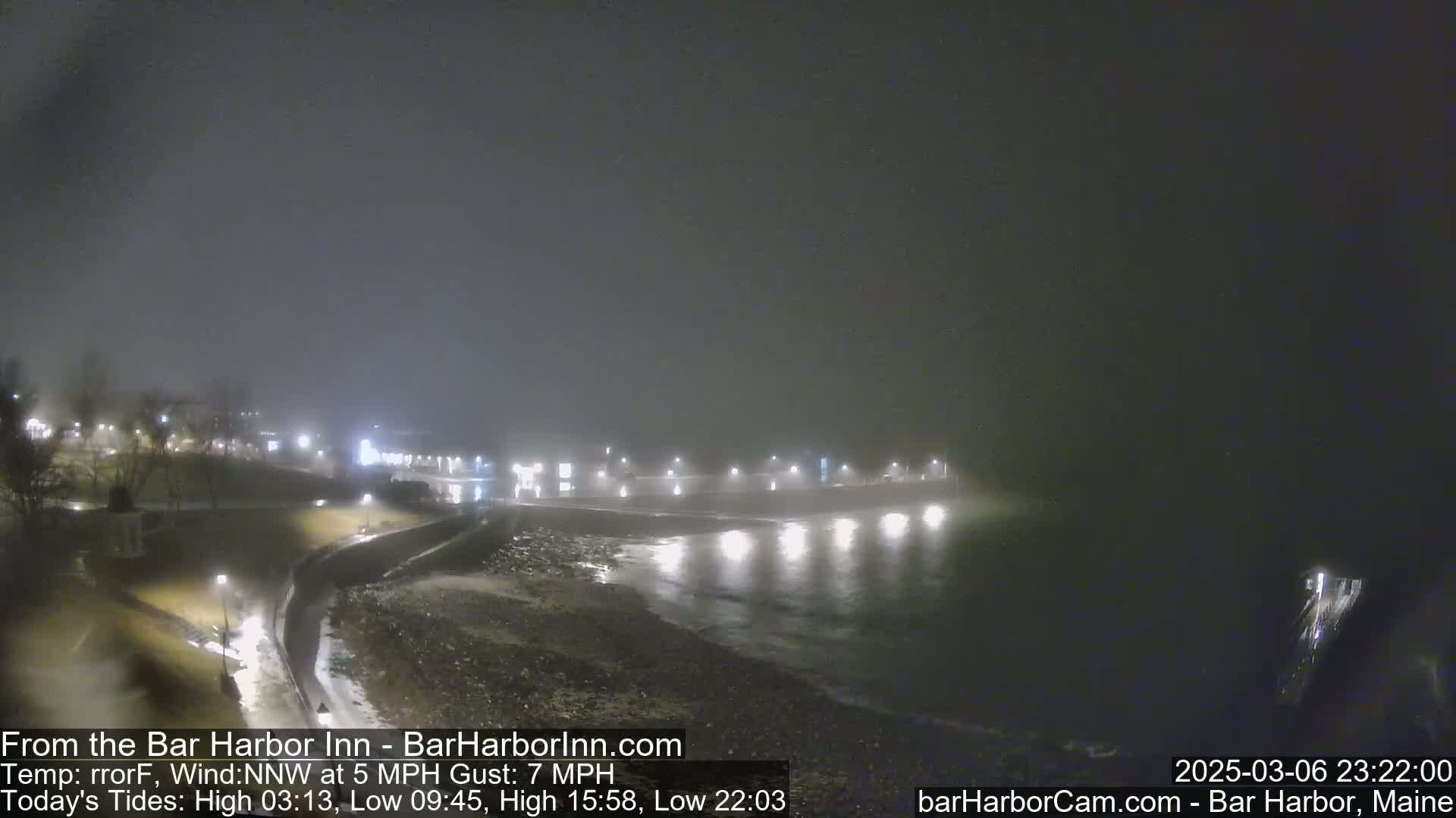 A foggy night scene shows a dark shoreline with lights illuminating a calm body of water and a distant town.